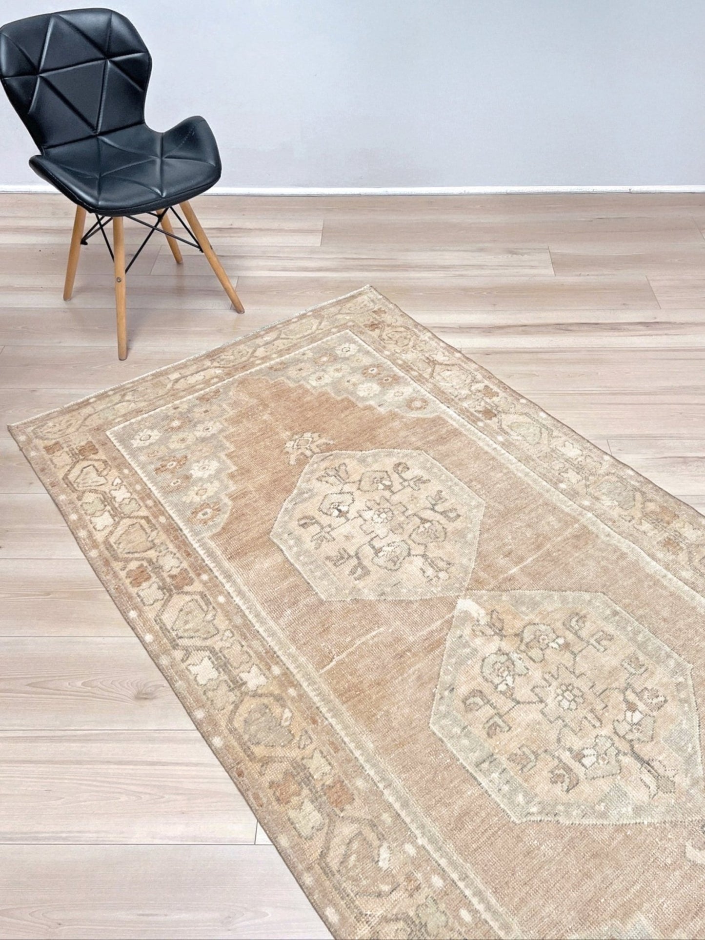 A rectangular area rug with a distressed traditional pattern in beige, tan, and cream, shown on a light wood floor next to a black modern chair.