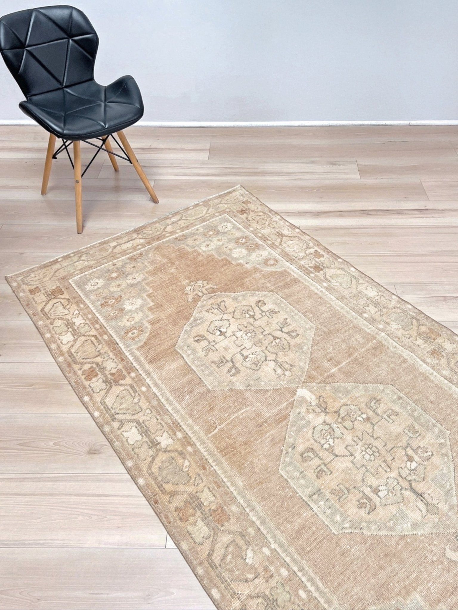 A rectangular area rug with a distressed traditional pattern in beige, tan, and cream, shown on a light wood floor next to a black modern chair.