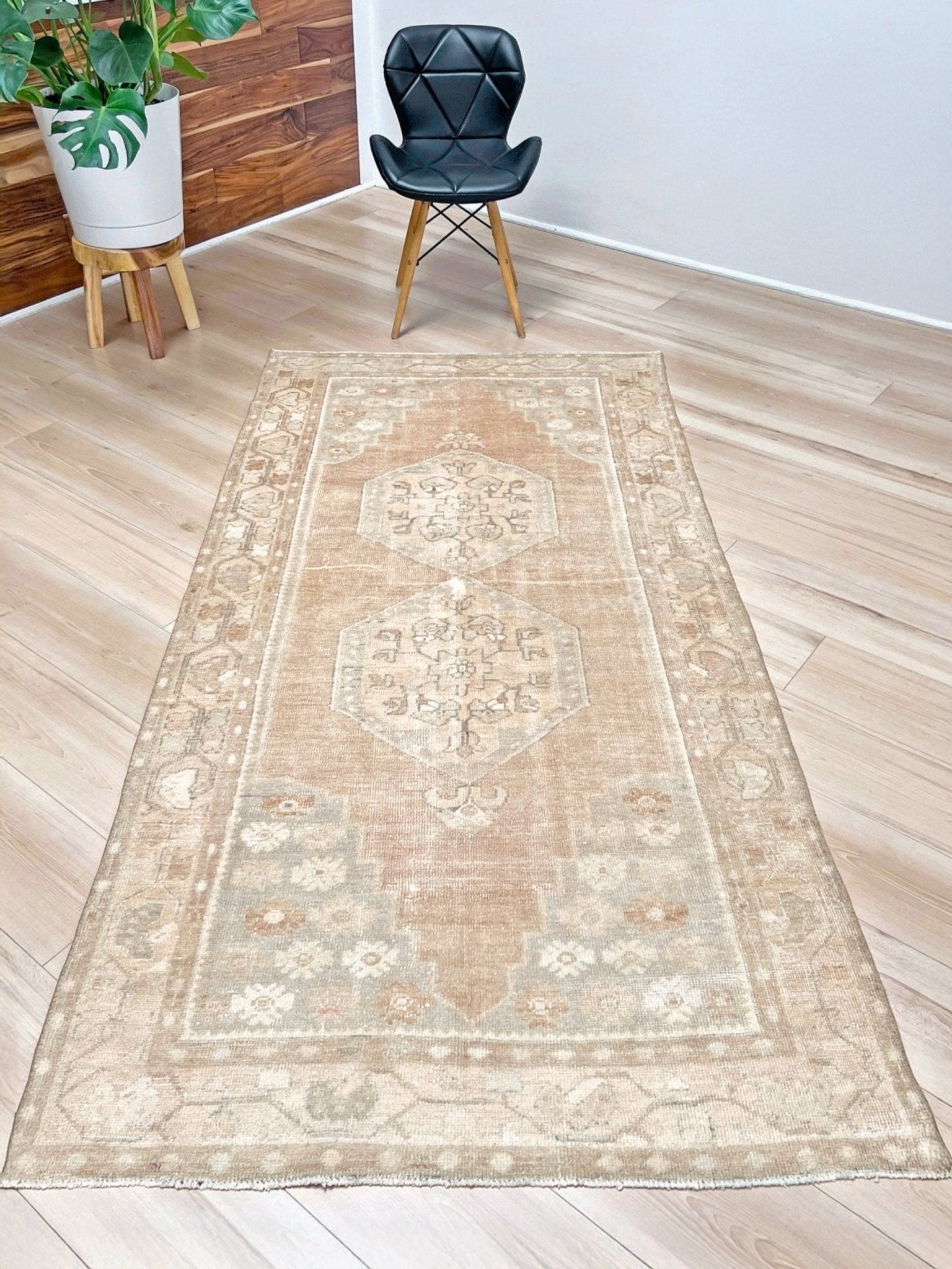 A rectangular area rug with a faded beige and light brown traditional medallion pattern, shown on a light wood floor in a room setting with a black chair and a potted plant.
