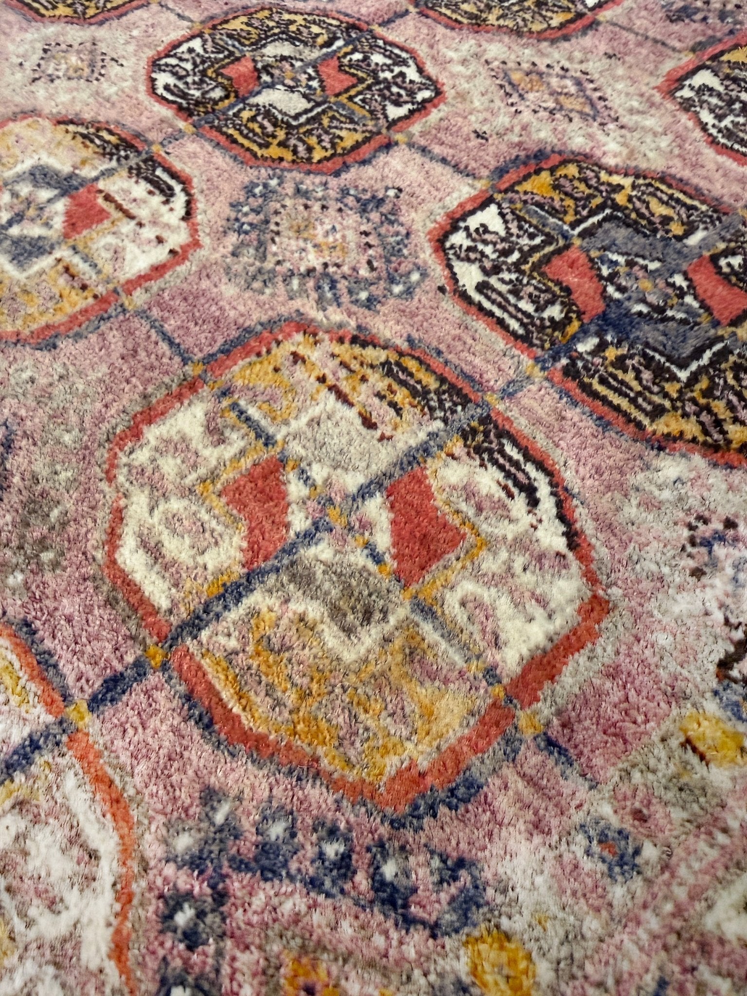 Close-up of a patterned rug with geometric designs in red, yellow, and black on a pink background. Earthy Muted Bukhara Turkmen rug  vintage handwoven wool carpet with tribal gul motifs and aged patina, 4x5 neutral area rug for entry under desk nursery, living room, bedroom, study, office.
