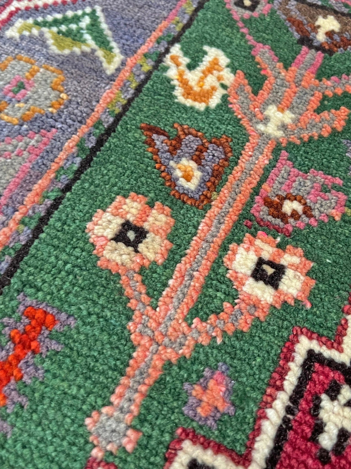 Close-up of a textured woven rug with a green field, featuring stylized floral motifs in pink, orange, cream, gray, purple, and black.