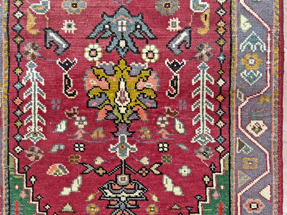 A close-up of a traditional handwoven rug with a deep red field, featuring a central floral medallion in muted green, yellow, and pink, surrounded by geometric borders in gray, green, and cream.