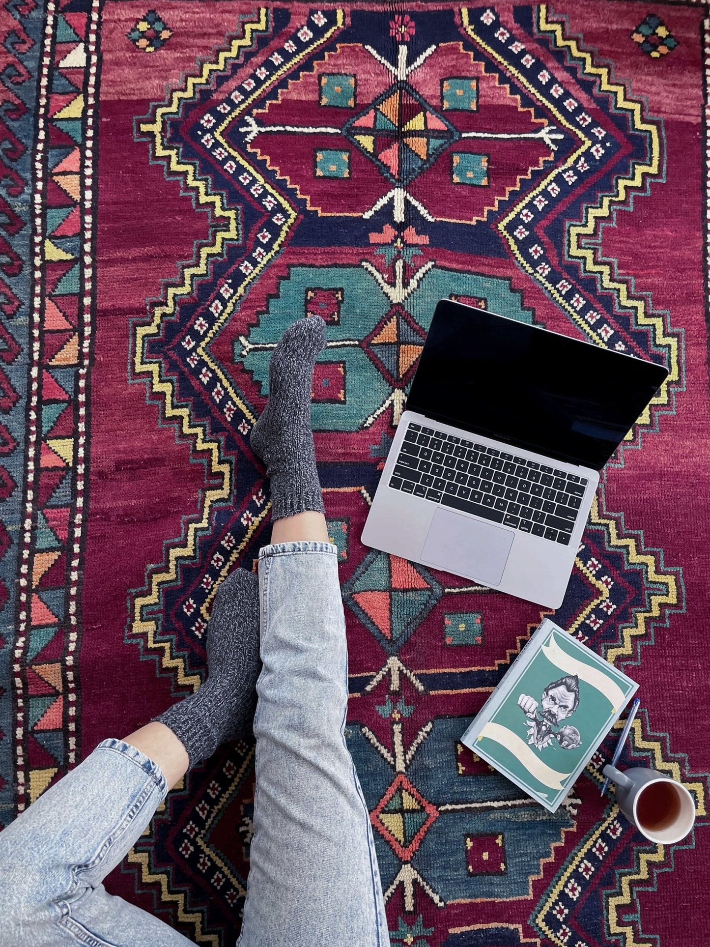Person using a laptop on a colorful patterned rug with a book and cup nearby. vintage kazak caucasian rug 6x9 burgundy field tribal geometric handmade wool 1940s antique style rug san francisco bay area rug shop indigo rugs