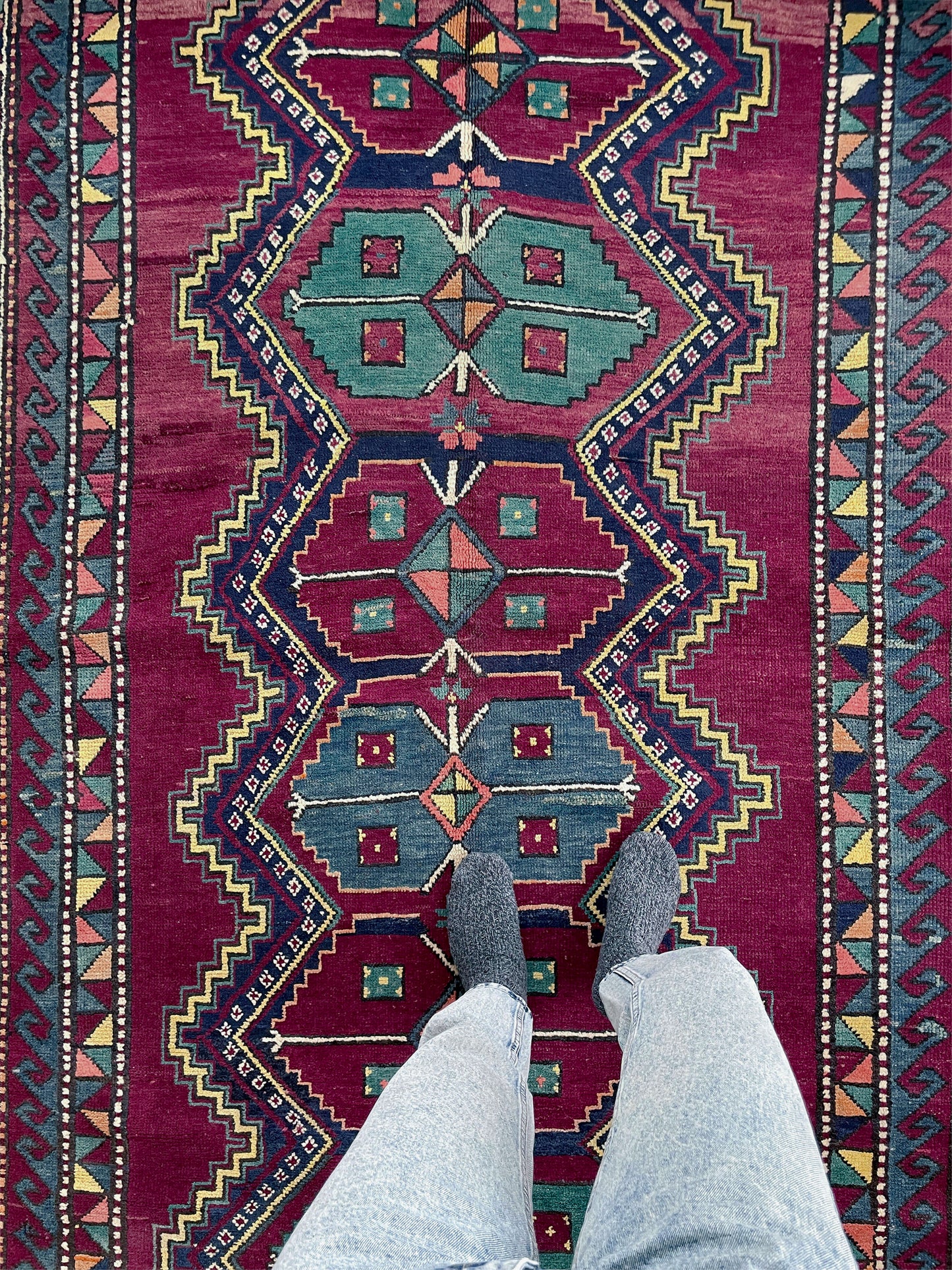 Person wearing shoes and light-colored pants standing on a vibrant, patterned rug. vintage kazak caucasian rug 6x9 burgundy field tribal geometric handmade wool 1940s antique style rug san francisco bay area rug shop indigo rugs
