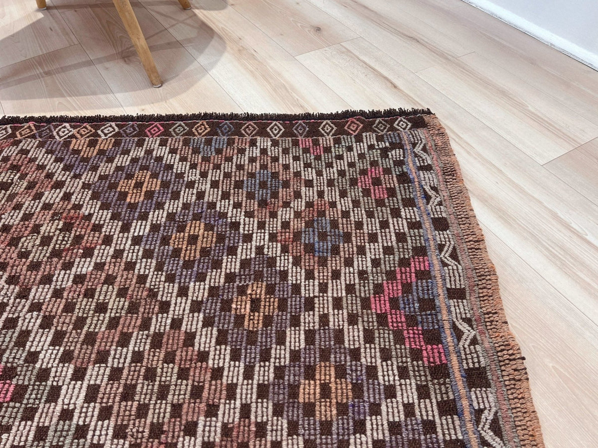 A close-up of a patterned area rug with a dark brown base and geometric motifs in beige, pink, and blue, laid out on a light wood floor.