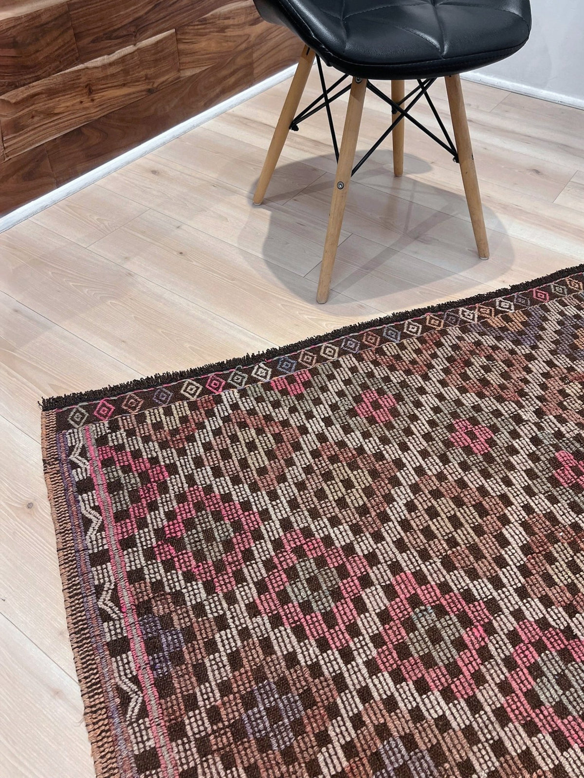 A close-up of a traditional kilim area rug with a geometric pattern in brown, pink, beige, and black, styled on a light wood floor next to a black chair with wooden legs.