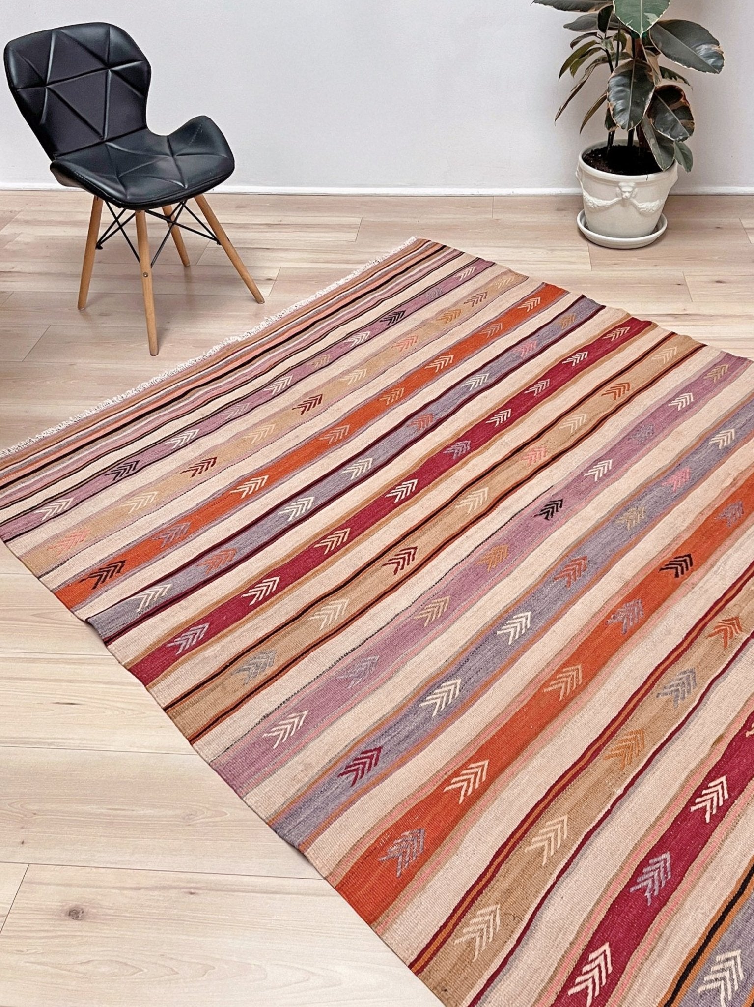 Striped area rug on a wooden floor with a chair and plant in the background. Vintage Anatolian kilim rug, 6x8 with horizontal arrow motifs in sun-washed tones.
