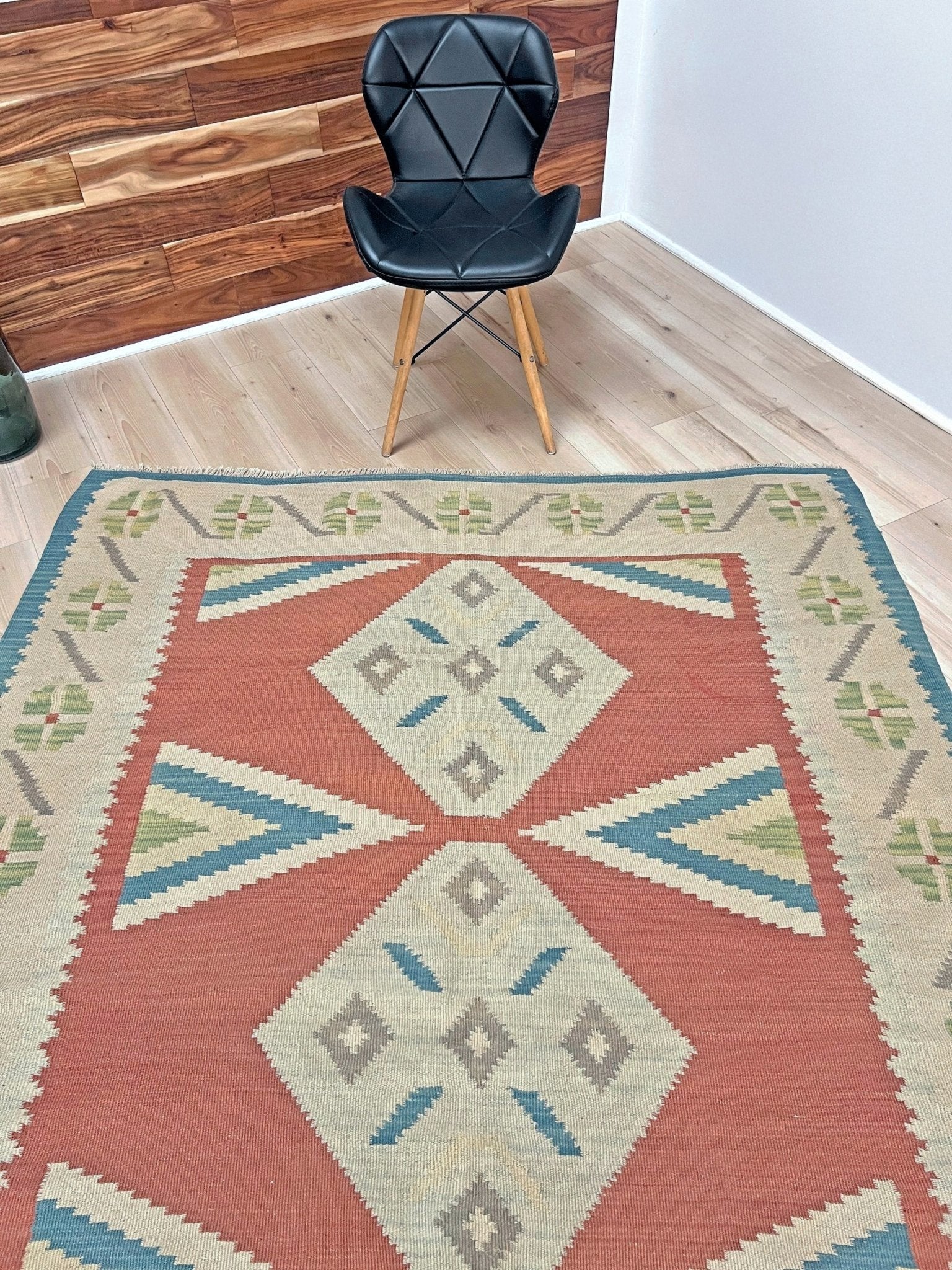 Patterned rug with geometric design on a wooden floor, black chair in the background. Vintage Turkish Malatya kilim rug with coral red field and diamond medallions, handwoven Anatolian flatweave 5x8 geometric wool rug