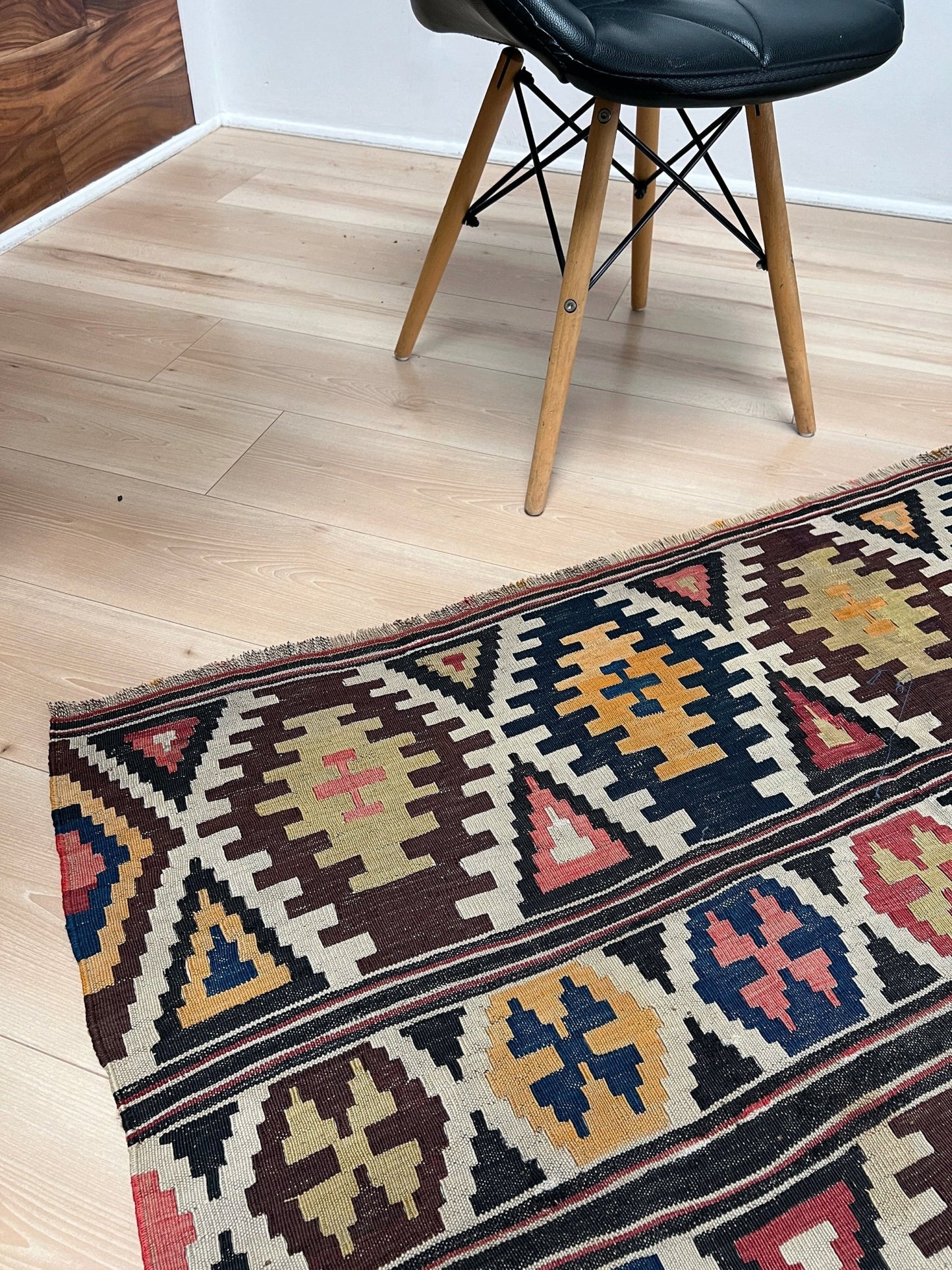 Patterned rug on a wooden floor with a black chair in the background. Vintage Caucasian Kazak tribal kilim rug with geometric medallions in beige, red, blue, and brown — handmade flatweave 5x8
