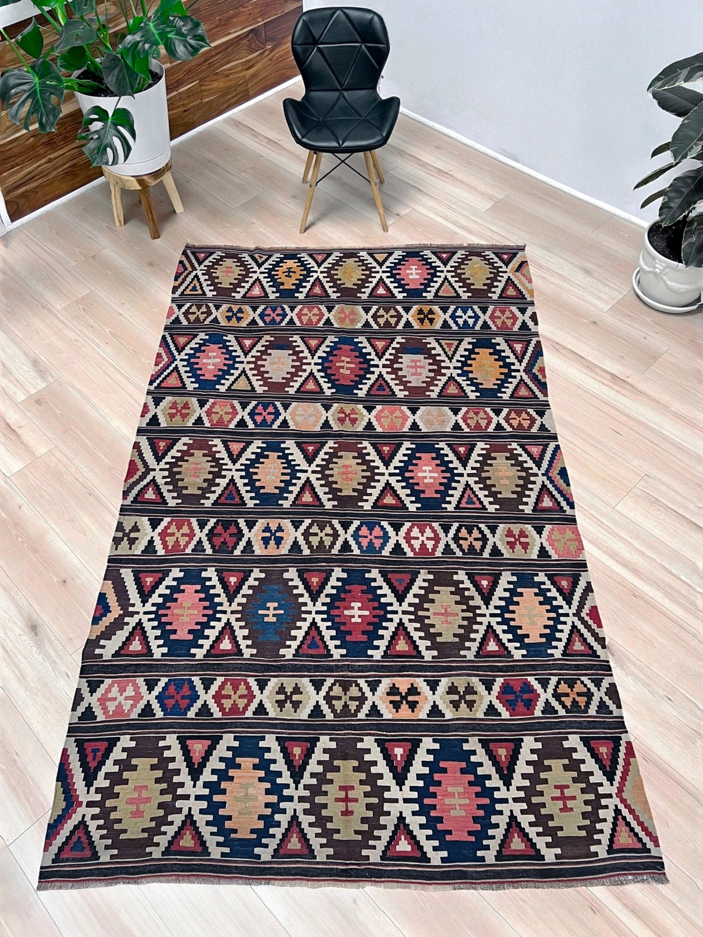 Patterned rug on a wooden floor with a chair and plants in the background. Vintage Caucasian Kazak tribal kilim rug with geometric medallions in beige, red, blue, and brown — handmade flatweave 5x8