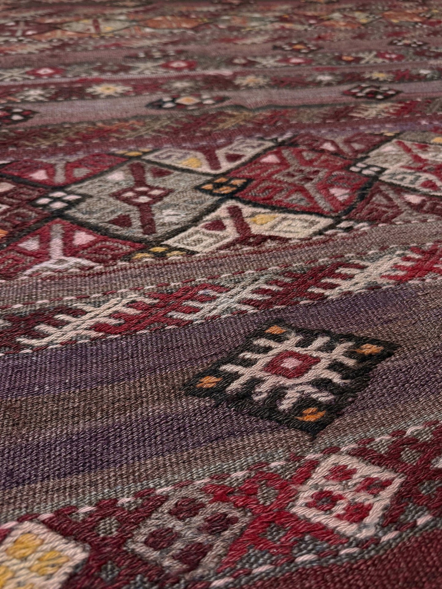Close-up of a Traditional patterned rug with geometric design on a plain background. 7x13 eggplant purple oxblood red Vintage 1940s Kurdish Kars kilim rug with tribal motifs for long living rooms, hallways, great rooms, and layered interiors. From Indigo Rugs, Burlingame CA.