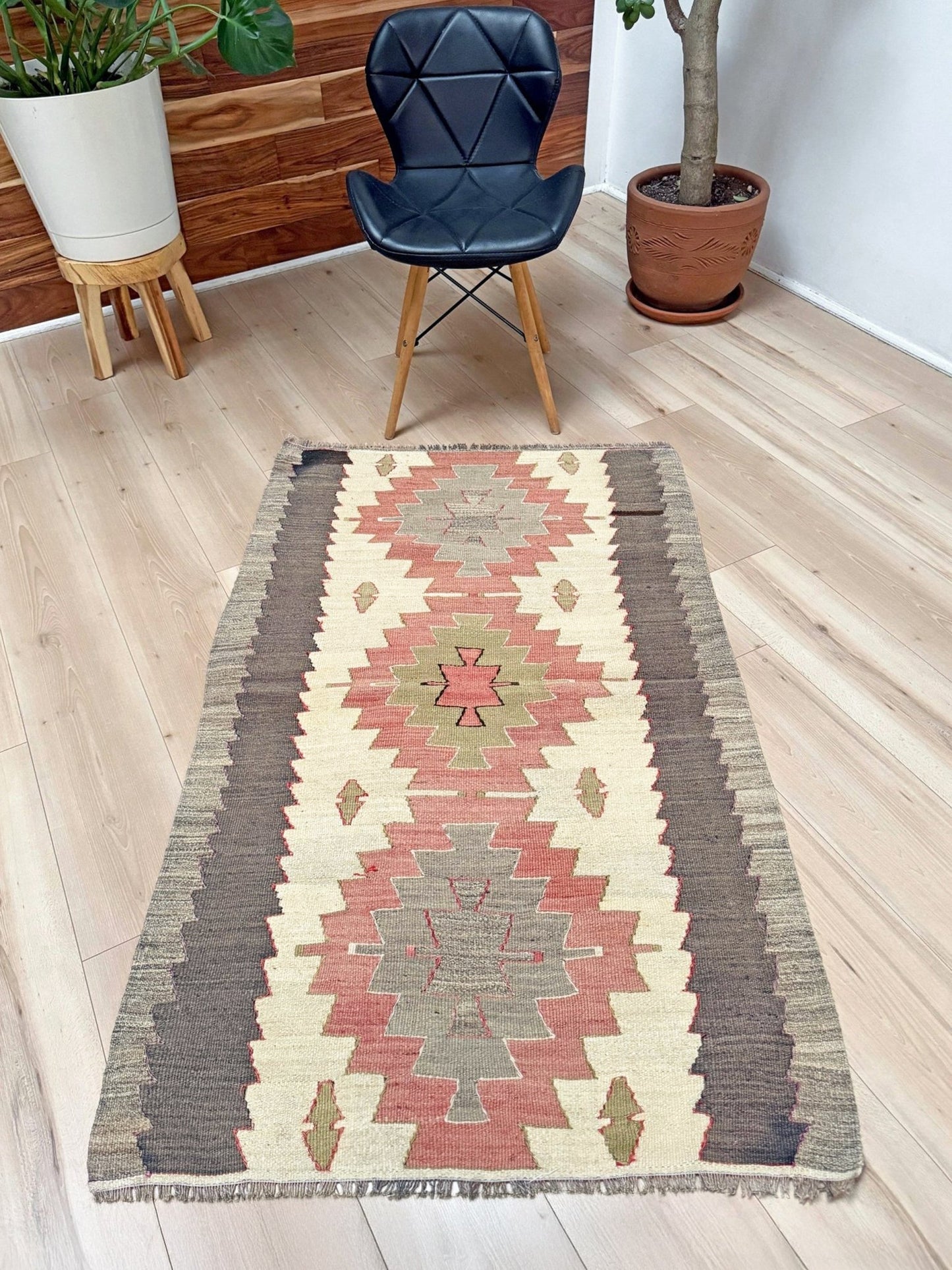 A rectangular woven area rug with a Southwestern geometric pattern in beige, terracotta, brown, and muted green, styled on a light wood floor in a room setting with a black chair and potted plants.