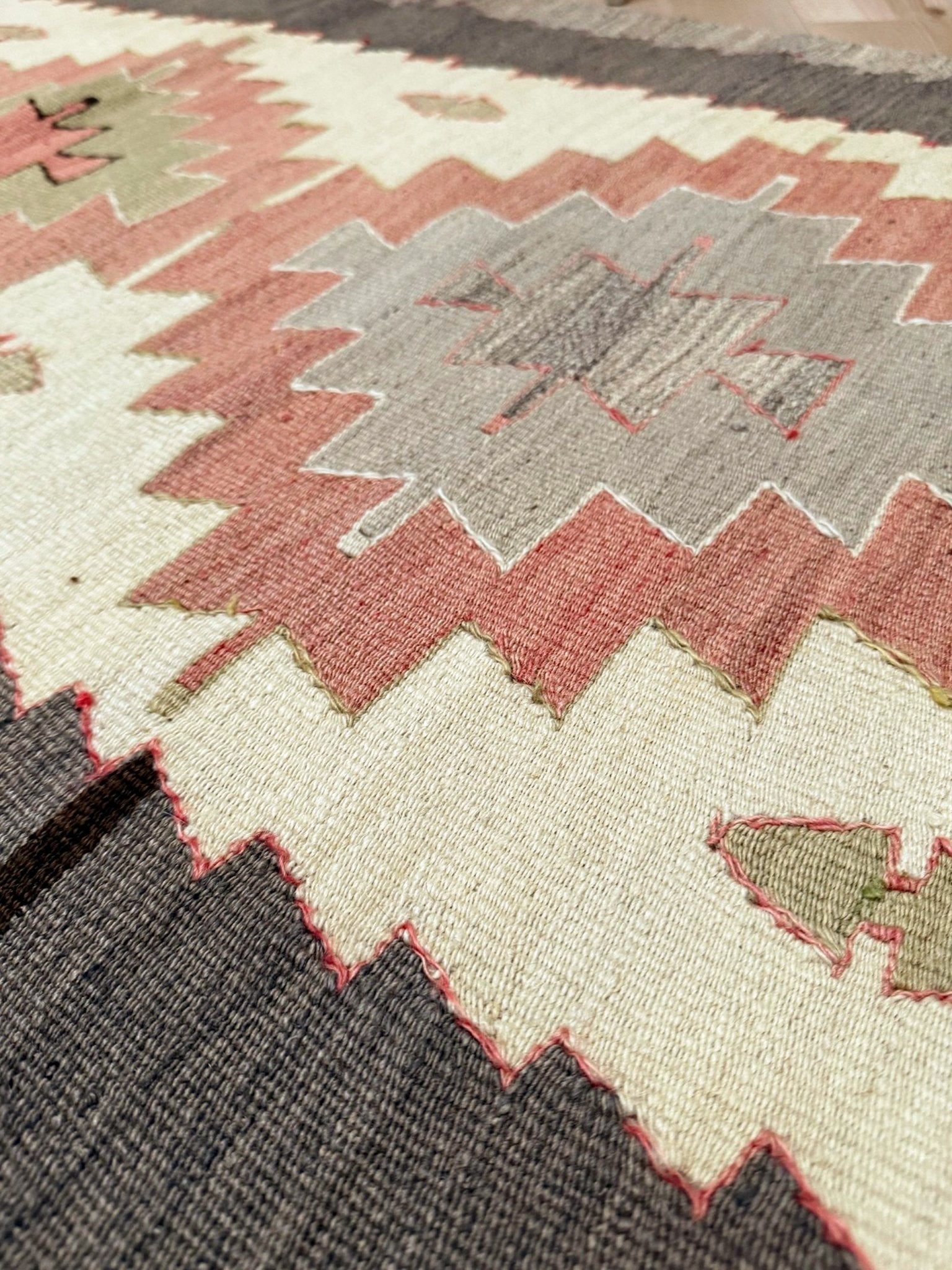 Close-up of a woven rug with a geometric pattern in cream, dusty pink, charcoal gray, and muted olive green.