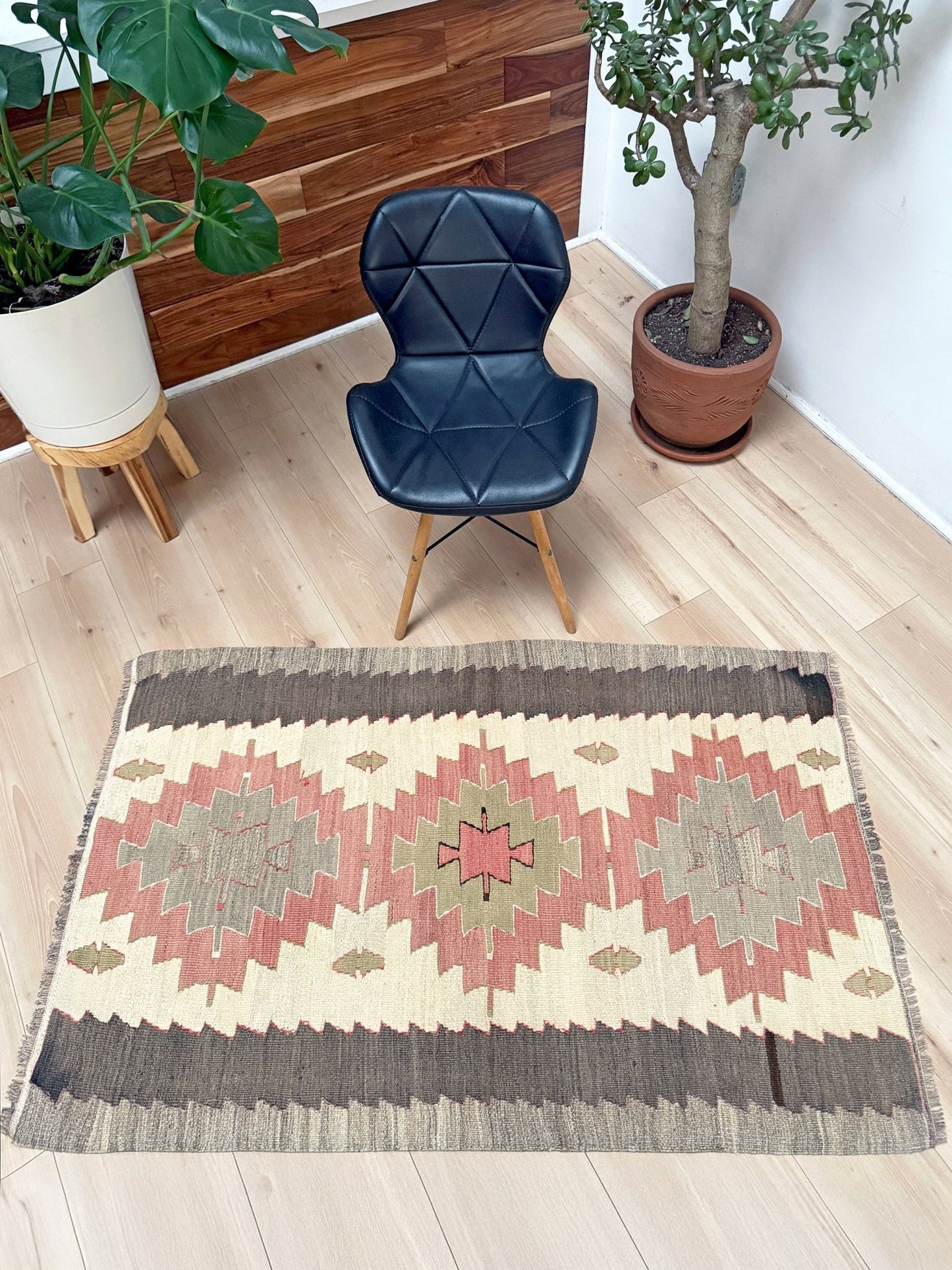 A rectangular area rug with a Southwestern geometric pattern in beige, brown, and dusty red, displayed on a light wood floor in a styled room with a black chair and potted plants.