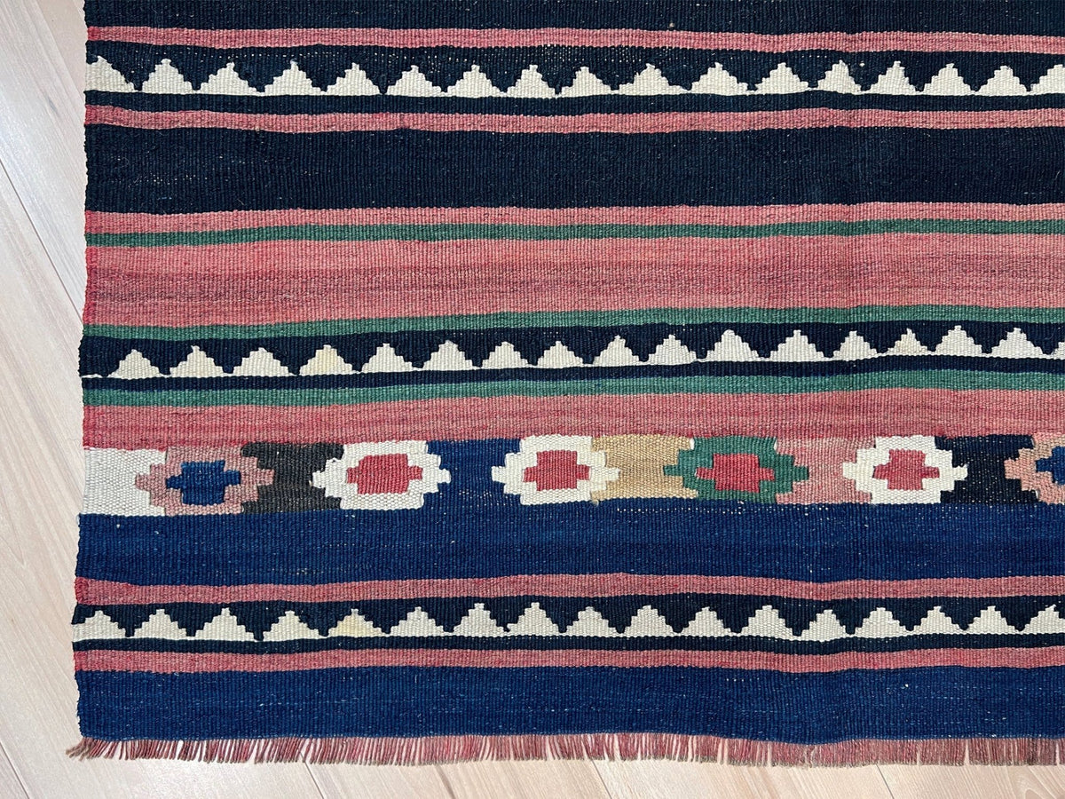 A close-up of a woven kilim rug with horizontal stripes and geometric patterns in navy blue, dusty pink, cream, green, and tan, laid on a light wood floor.