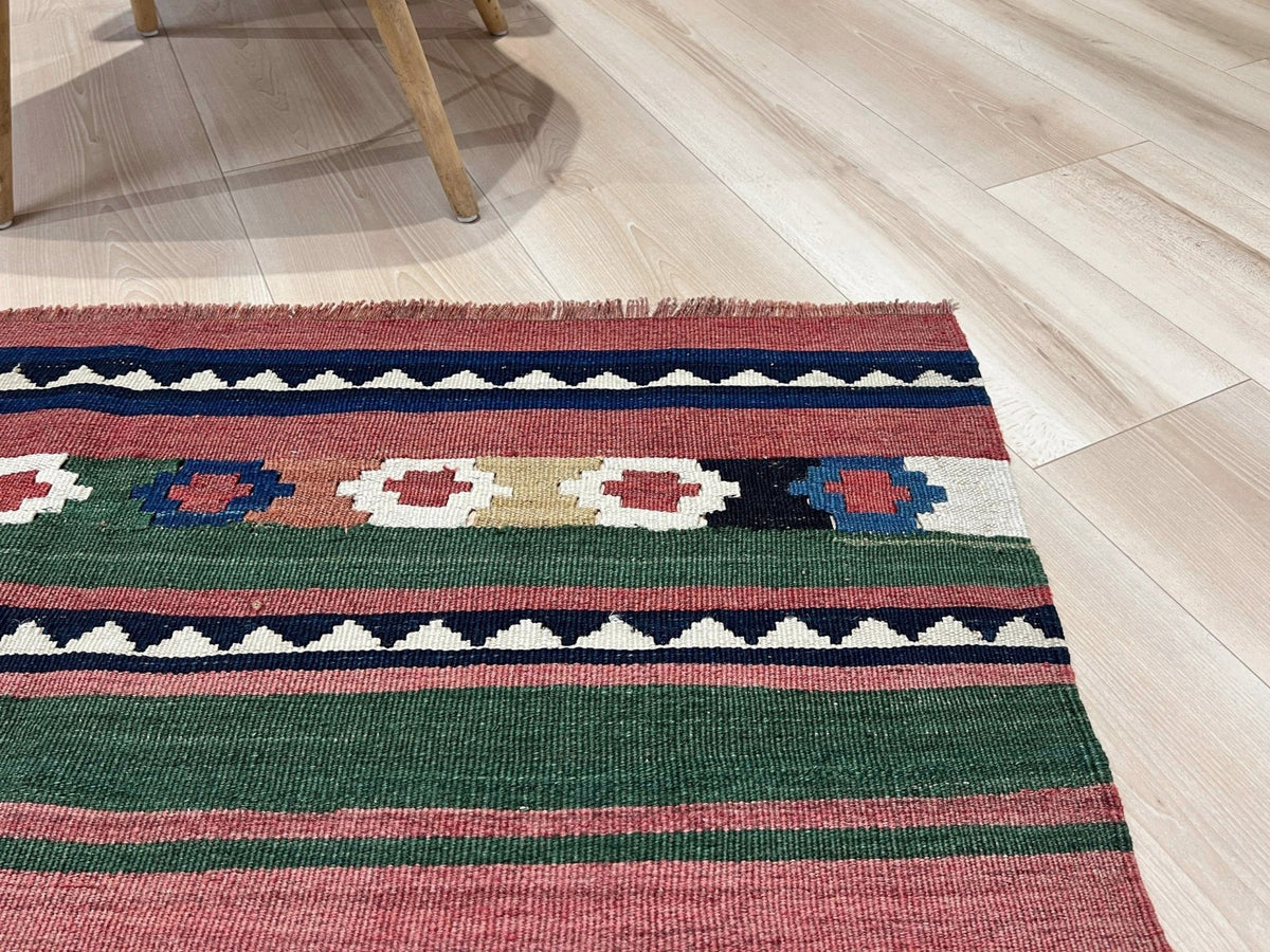 A close-up of a woven kilim rug with geometric patterns in green, pink, navy, cream, and beige, laid out on a light wood floor.