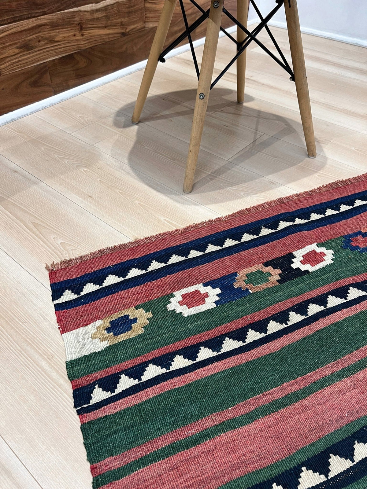 A close-up of a woven kilim-style area rug with geometric stripes in green, red, navy, cream, and tan, shown on a light wood floor near the legs of a wooden chair.