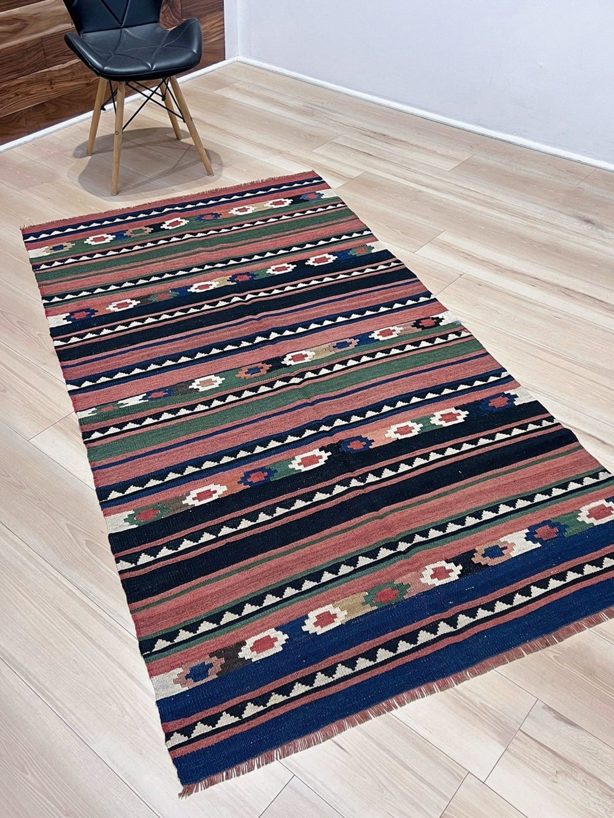A rectangular kilim runner rug with a colorful geometric and floral pattern, displayed on a light wood floor next to a black modern chair.