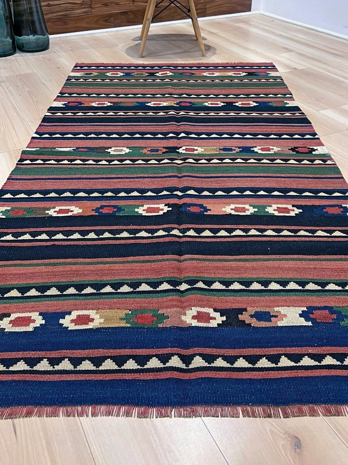 A rectangular kilim area rug with a colorful geometric tribal pattern, laid out on a light wood floor in a room setting.