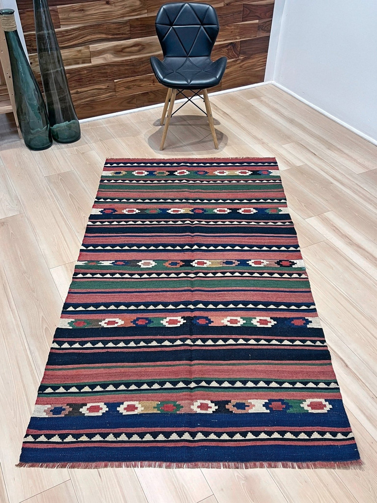 A long rectangular kilim runner rug with a colorful geometric pattern, laid out on a light wood floor in a room setting with a black chair and wooden wall paneling in the background.