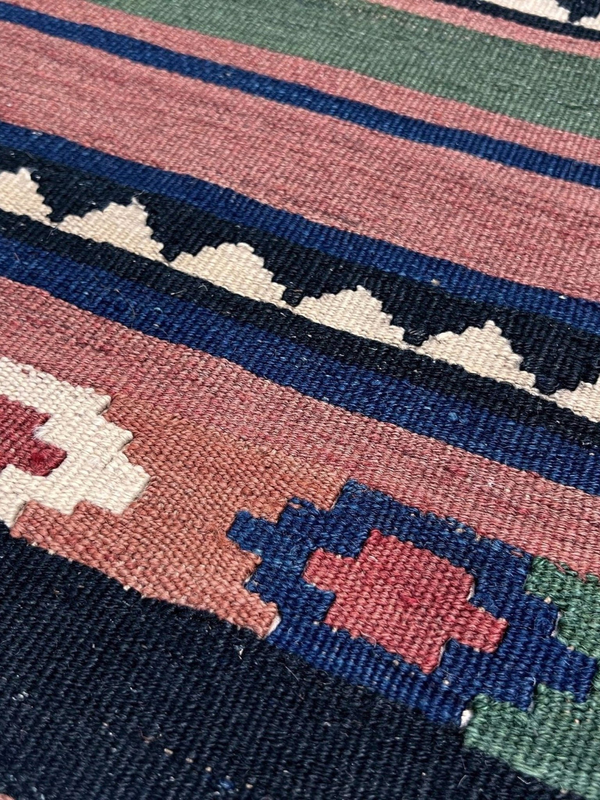 Close-up of a woven kilim rug showing geometric stripes and motifs in navy blue, dusty pink, cream, green, and red.