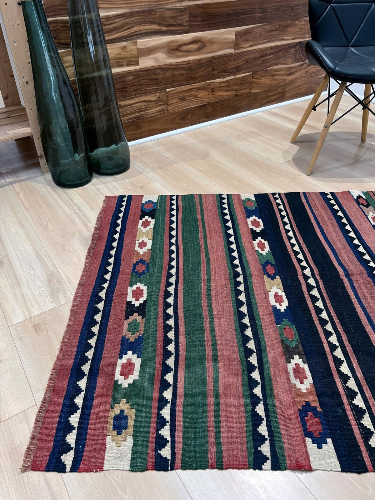A rectangular kilim-style area rug with vertical stripes and geometric motifs in red, green, navy, cream, and beige, shown on a light wood floor in a modern room setting.