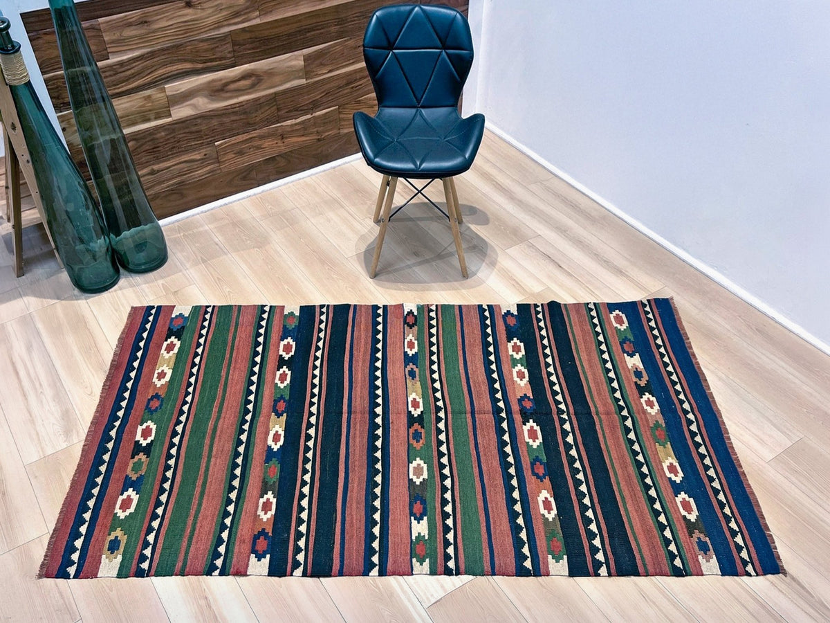 A rectangular kilim-style area rug with vertical stripes and geometric motifs in navy, red, green, beige, and brown, displayed on a light wood floor in a room setting with a blue chair and glass vases.