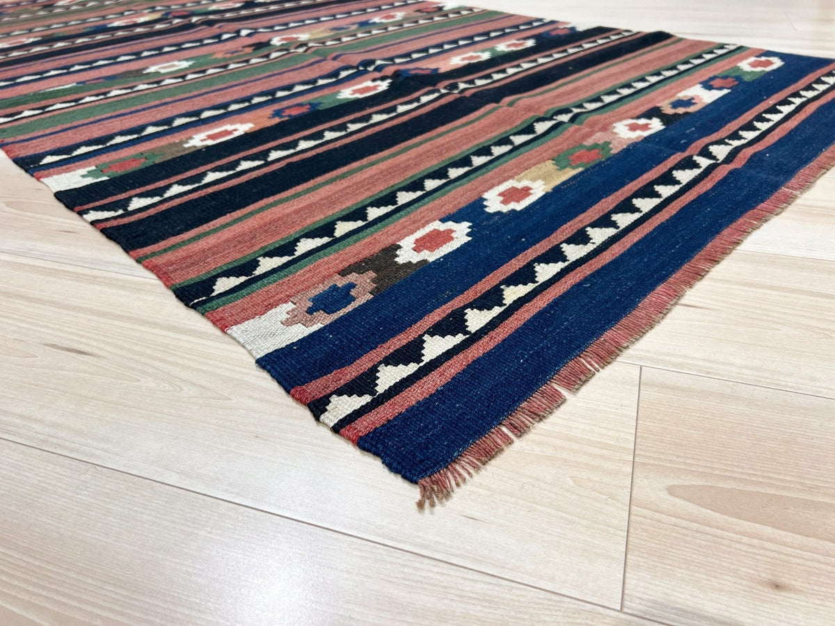 A square woven kilim rug with a geometric pattern, shown partially unrolled on a light-colored wooden floor. The rug features stripes of navy blue, terracotta, cream, and green with a fringed edge.