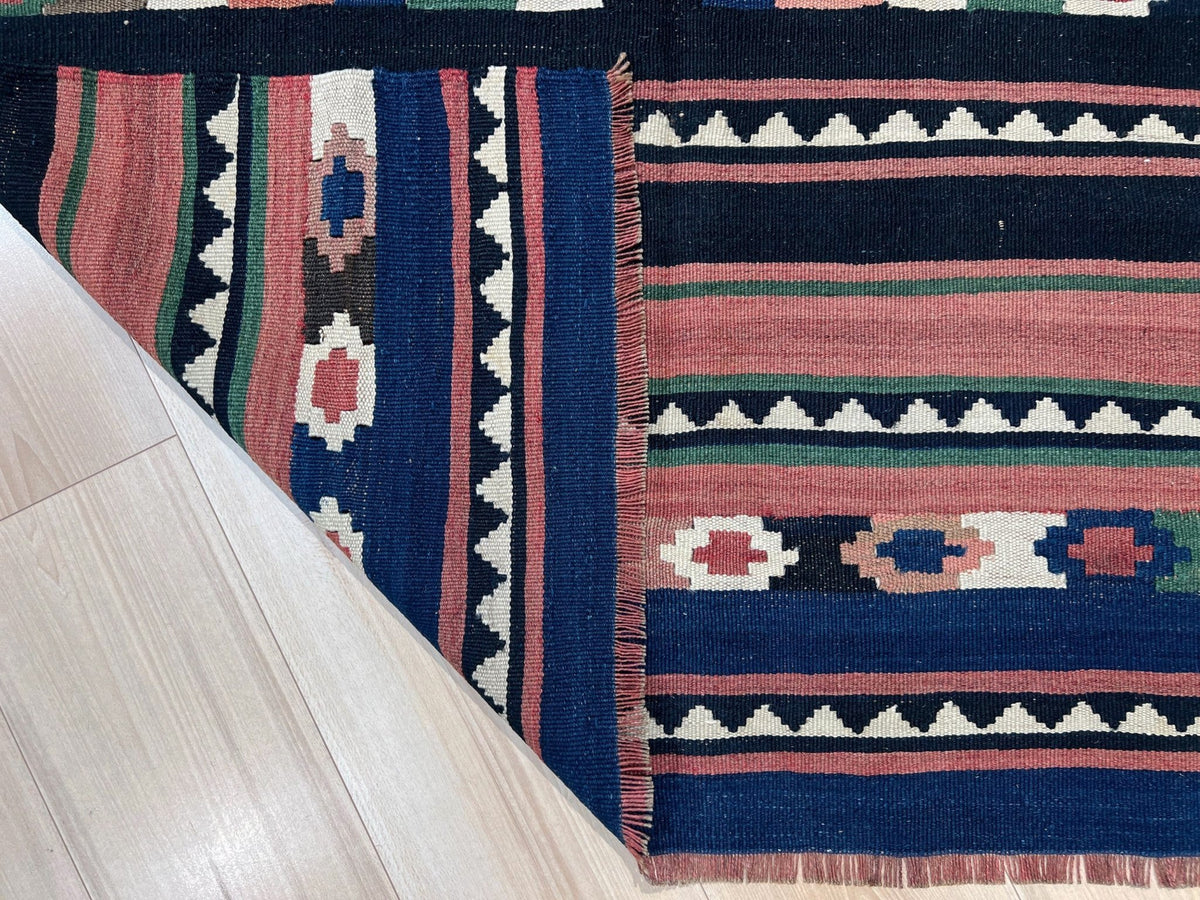 A close-up of a woven kilim rug with geometric patterns in navy blue, dusty pink, cream, and green, shown on a light wood floor with one corner folded over.