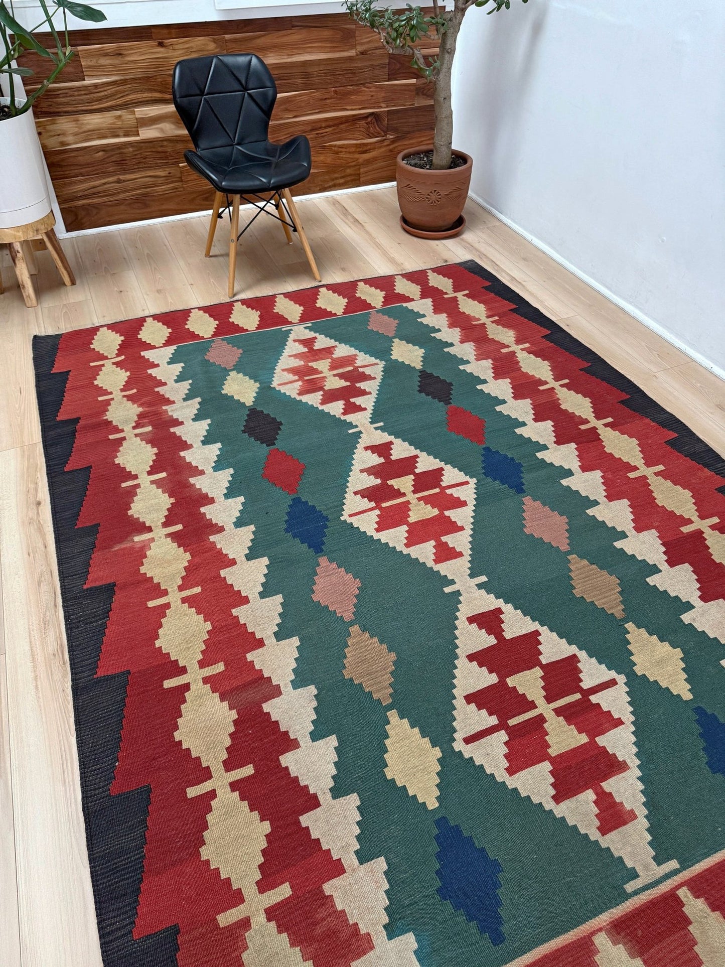 A rectangular area rug with a Southwestern geometric pattern in red, green, beige, blue, and black, styled on a light wood floor in a room with a black chair and potted plants.