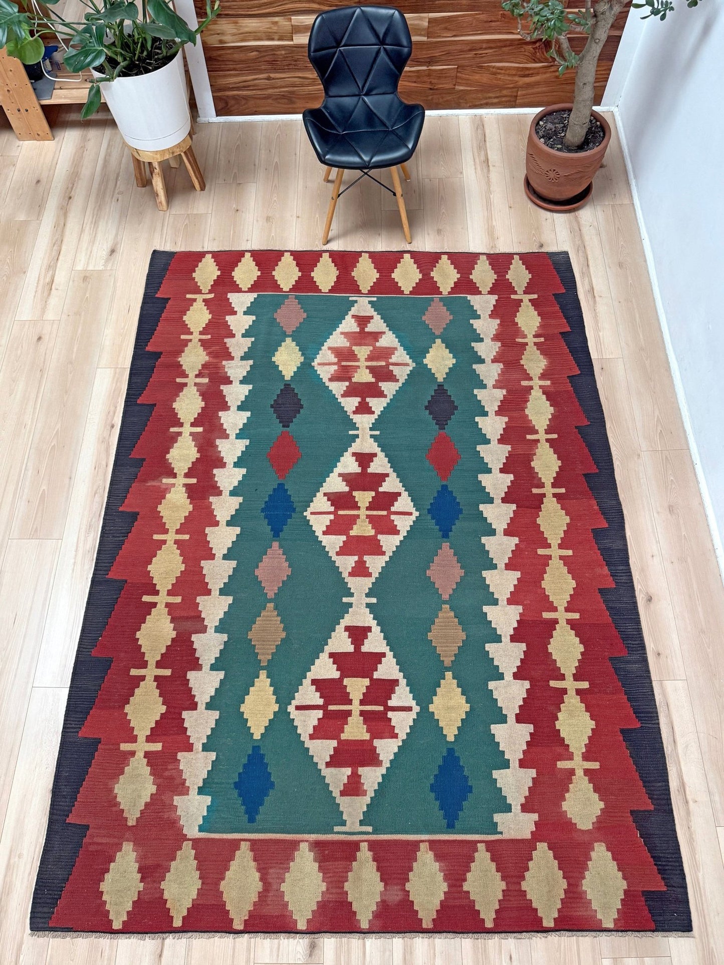 A rectangular area rug with a Southwestern geometric pattern in red, teal, beige, and blue, shown on a light wood floor in a styled room with a black chair and potted plants.