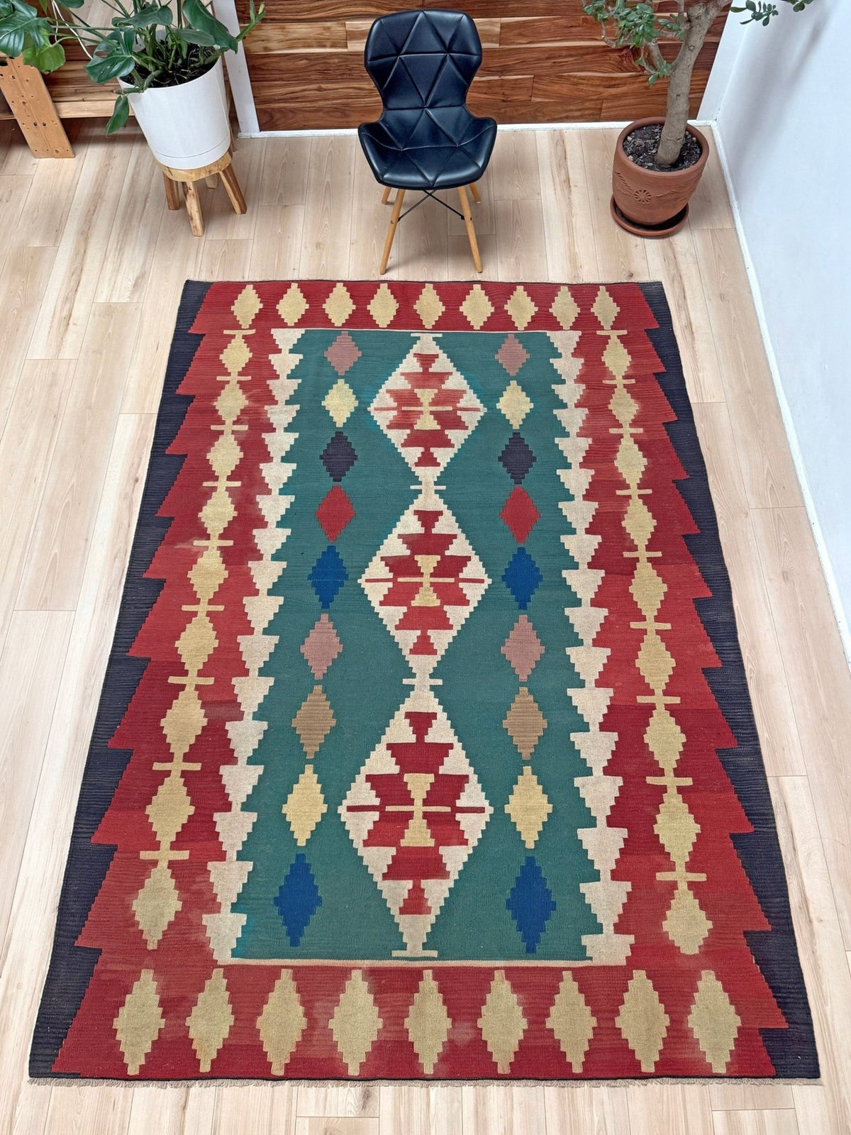 A rectangular Southwestern-style area rug with a geometric diamond and zigzag pattern in red, teal green, beige, blue, and dark brown, displayed on a light wood floor in a room setting with a black chair and potted plants.
Vibrant Denizli vintage Turkish kilim rug with Southwestern geometric pattern, handmade wool flatweave 6x8 from Indigo Rugs Burlingame Bay Area.