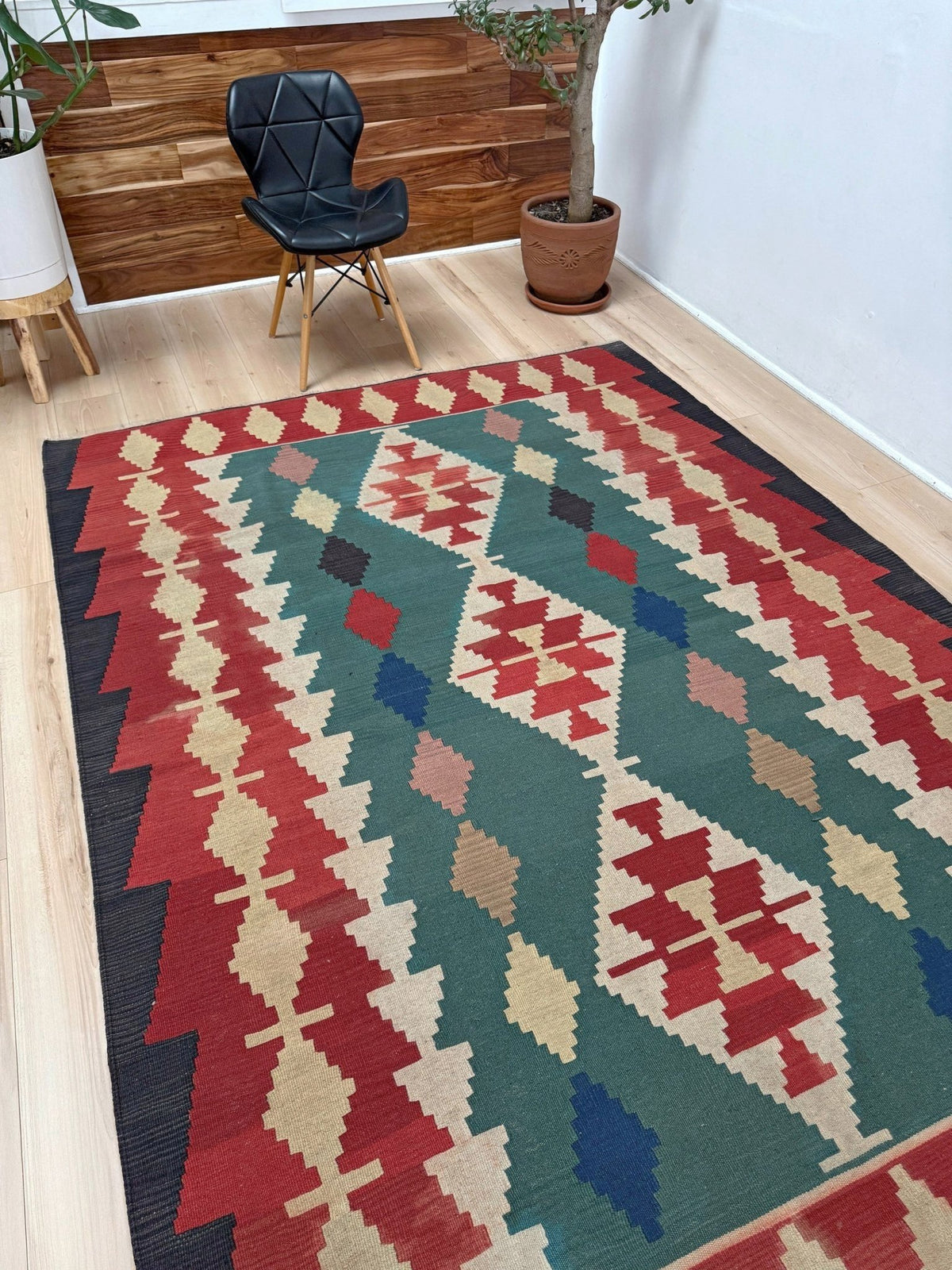 A rectangular area rug with a Southwestern geometric pattern in red, green, beige, blue, and black, styled on a light wood floor in a room with a black chair and potted plants.