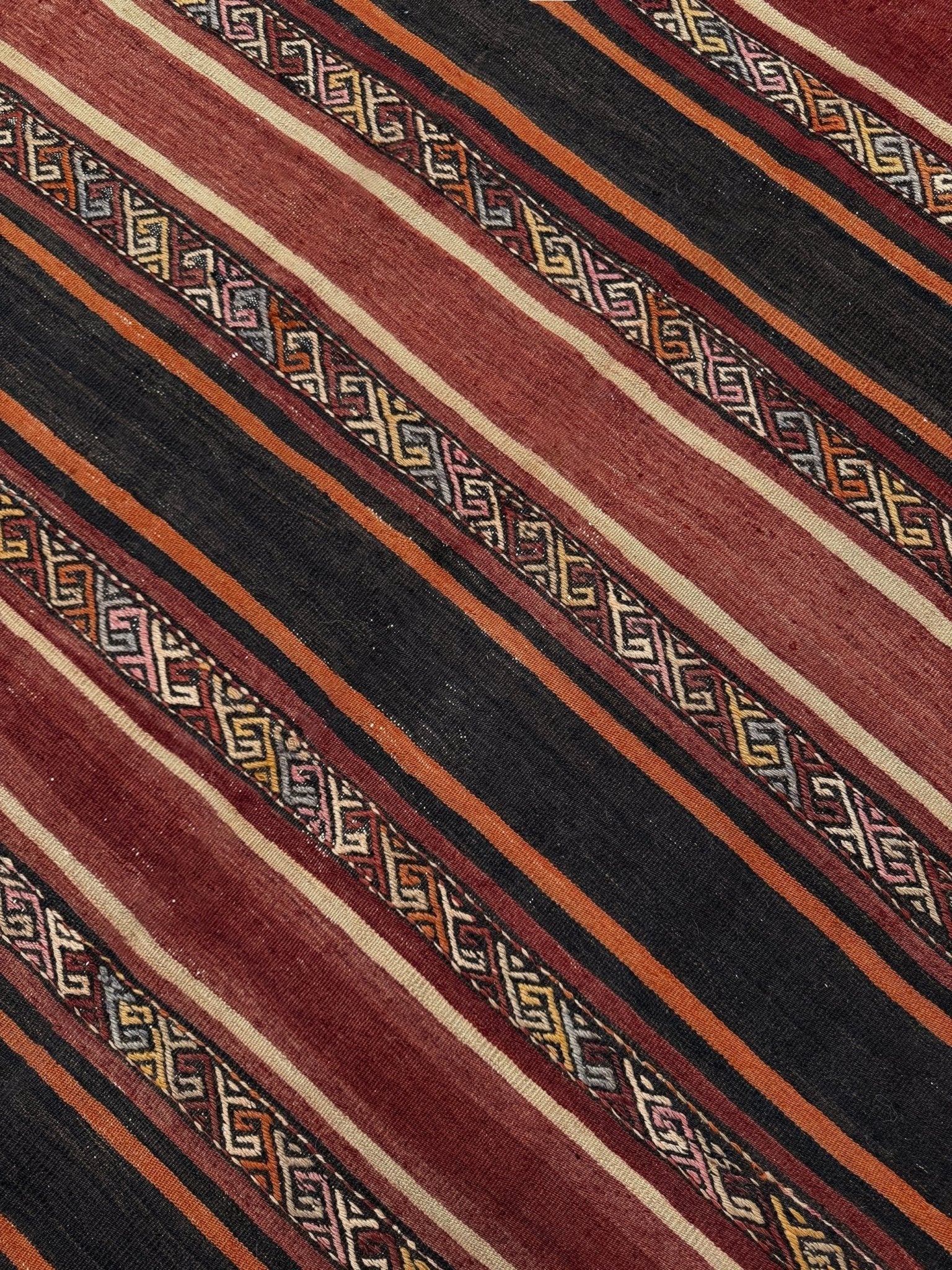 Close-up of a patterned fabric with red, black, and gold stripes. Earthy Large vintage Turkish kilim rug with horizontal tribal stripes in madder red black and earth tones, handmade wool flatweave.