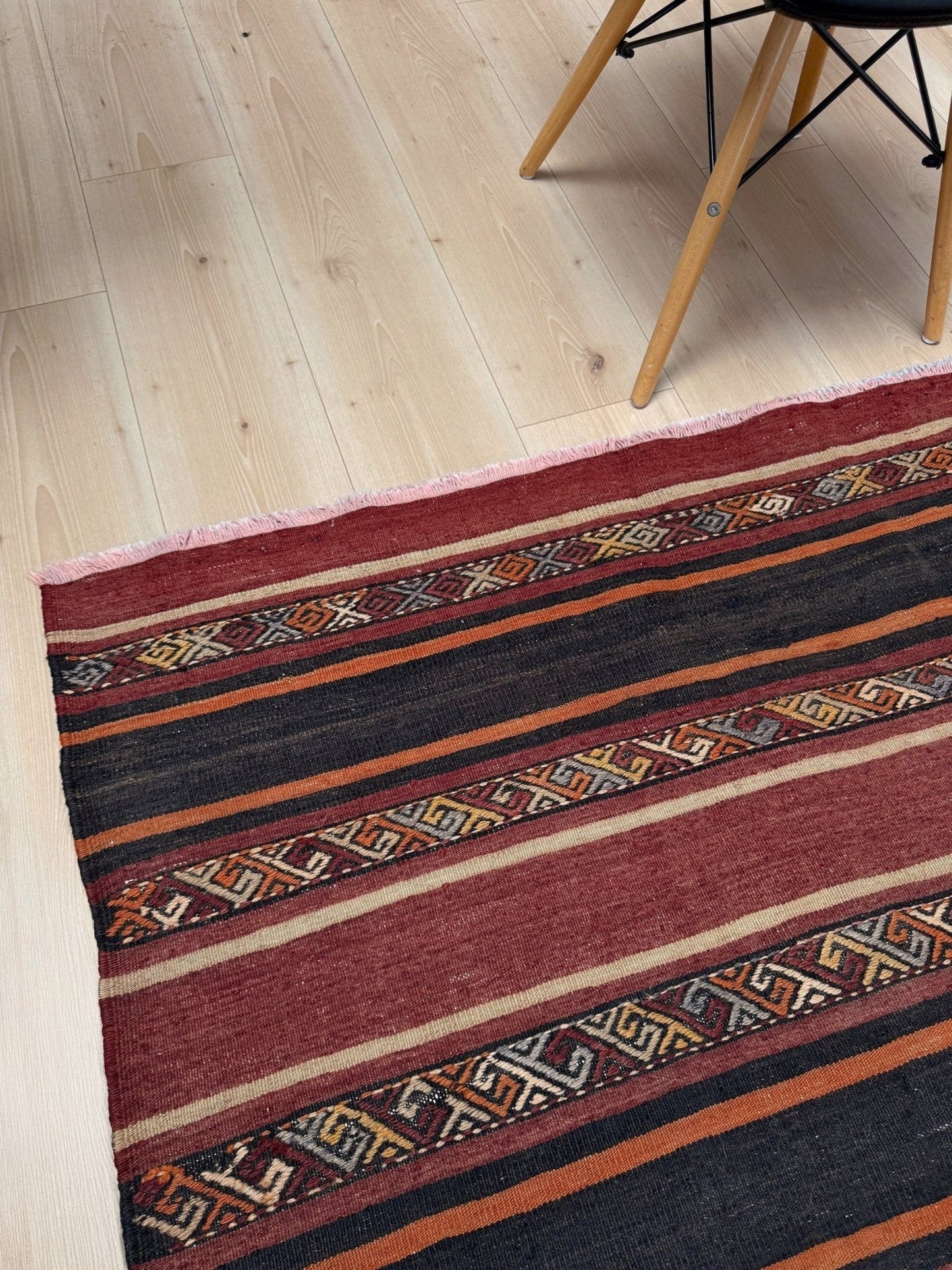 Striped patterned rug on a wooden floor with a chair partially visible. Earthy Large vintage Turkish kilim rug with horizontal tribal stripes in madder red black and earth tones, handmade wool flatweave.