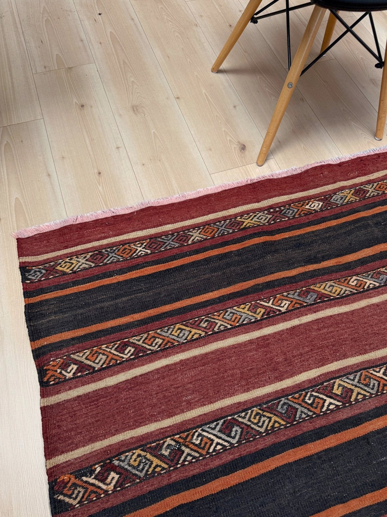 Striped patterned rug on a wooden floor with a chair partially visible. Earthy Large vintage Turkish kilim rug with horizontal tribal stripes in madder red black and earth tones, handmade wool flatweave.