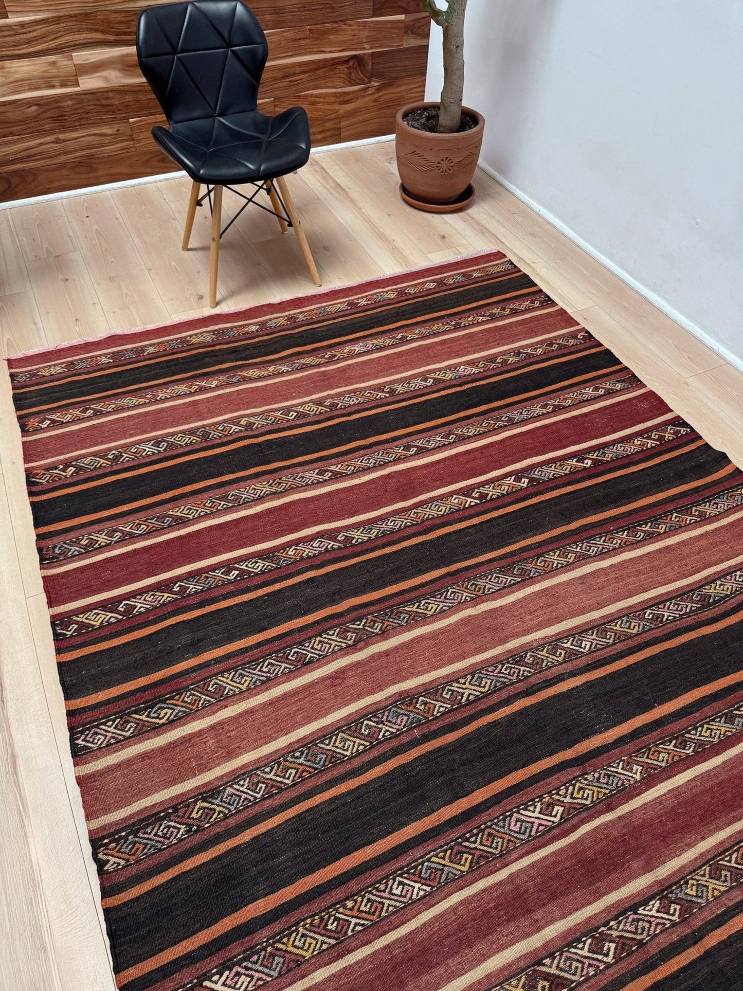 Striped red and black rug on a wooden floor with a chair and plant in the background. Earthy Large 6x9 vintage Turkish kilim rug with horizontal tribal stripes in madder red black and earth tones, handmade wool flatweave.