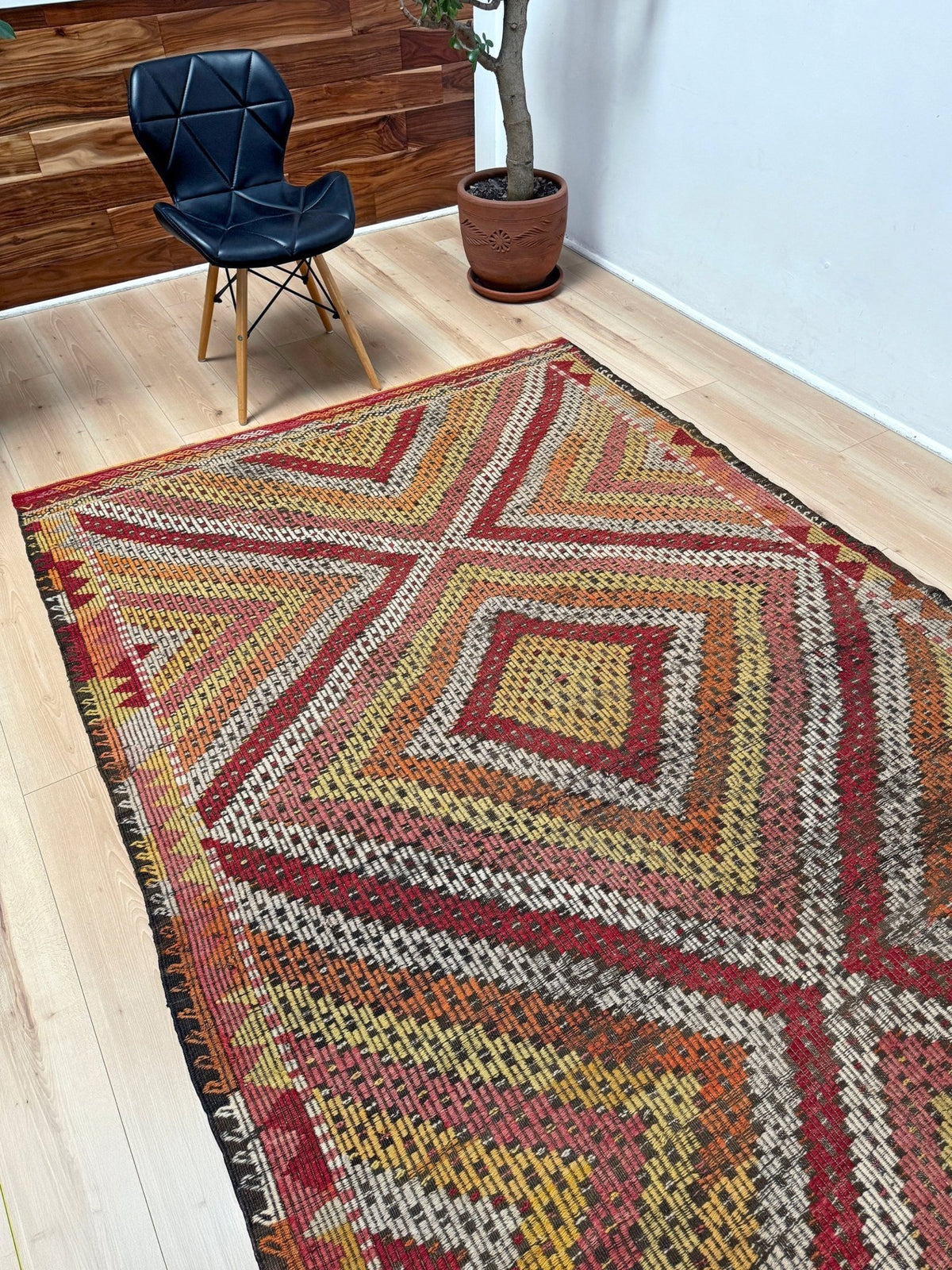 A large kilim area rug with a geometric diamond pattern in red, orange, yellow, and beige, styled on a light wood floor in a room with a black leather chair and a potted plant.  6x9 Vintage Turkish cicim kilim rug with bold diamond pattern, handmade Turkish embroidered wool flatweave from Indigo Rugs Burlingame Bay Area.