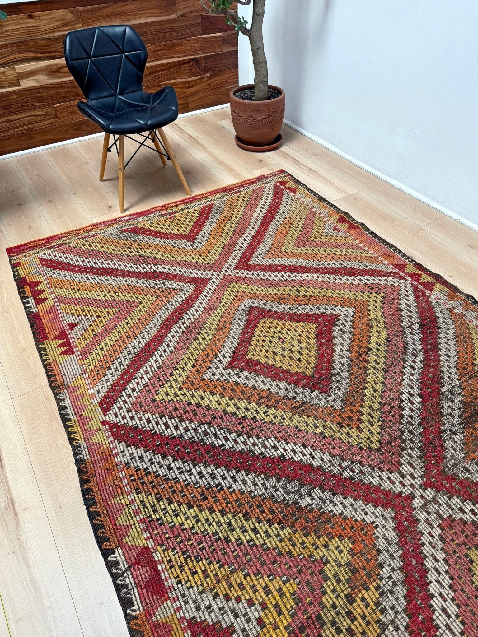 A large kilim area rug with a geometric diamond pattern in red, orange, yellow, and beige, styled on a light wood floor in a room with a black leather chair and a potted plant.  6x9 Vintage Turkish cicim kilim rug with bold diamond pattern, handmade Turkish embroidered wool flatweave from Indigo Rugs Burlingame Bay Area.