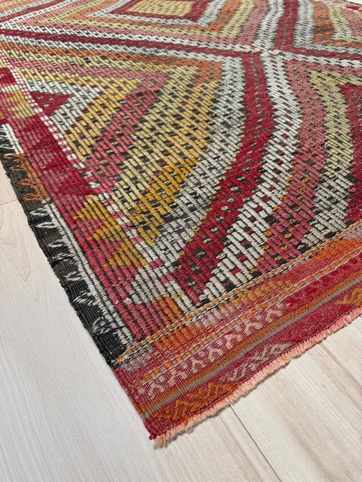 A close-up of a woven kilim rug corner with geometric patterns in red, yellow, orange, black, and cream, laid on a light wood floor.