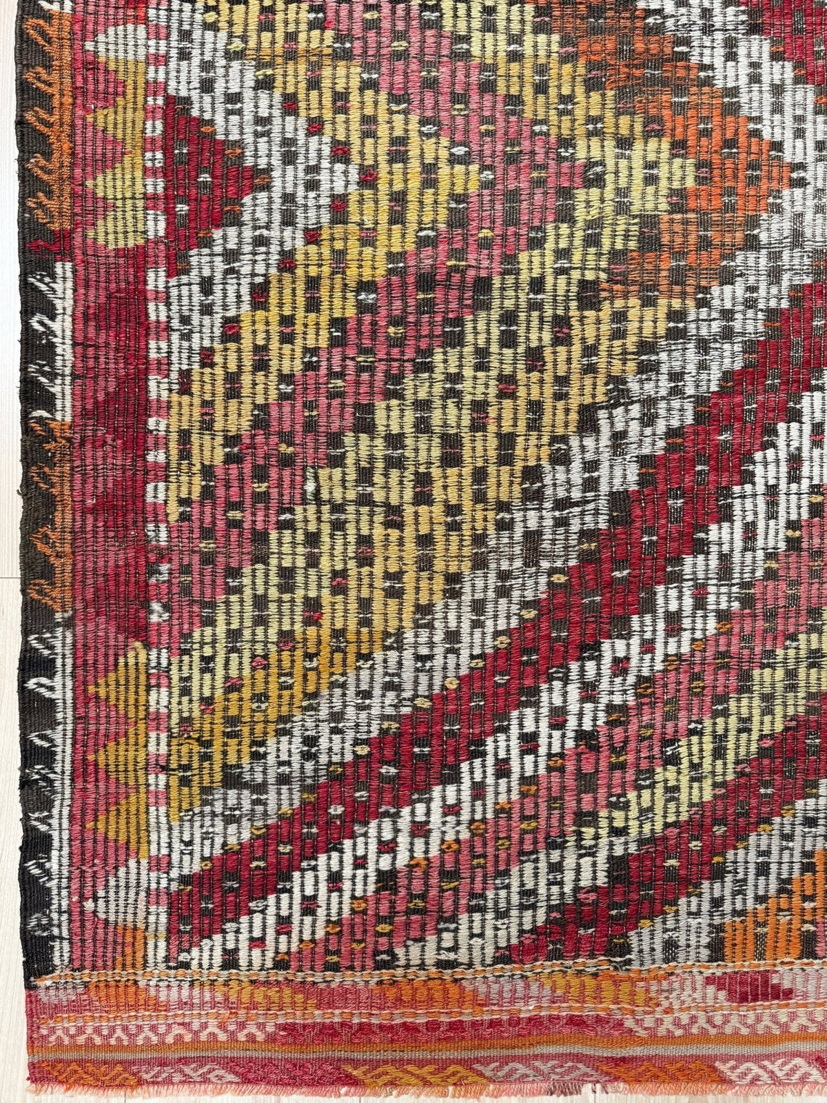 A close-up of a woven textile or rug showing a geometric pattern with diagonal bands in red, pink, yellow, black, white, and orange, plus a fringed edge and patterned border.  6x9 Vintage Turkish cicim kilim rug with bold diamond pattern, handmade Turkish embroidered wool flatweave from Indigo Rugs Burlingame Bay Area.