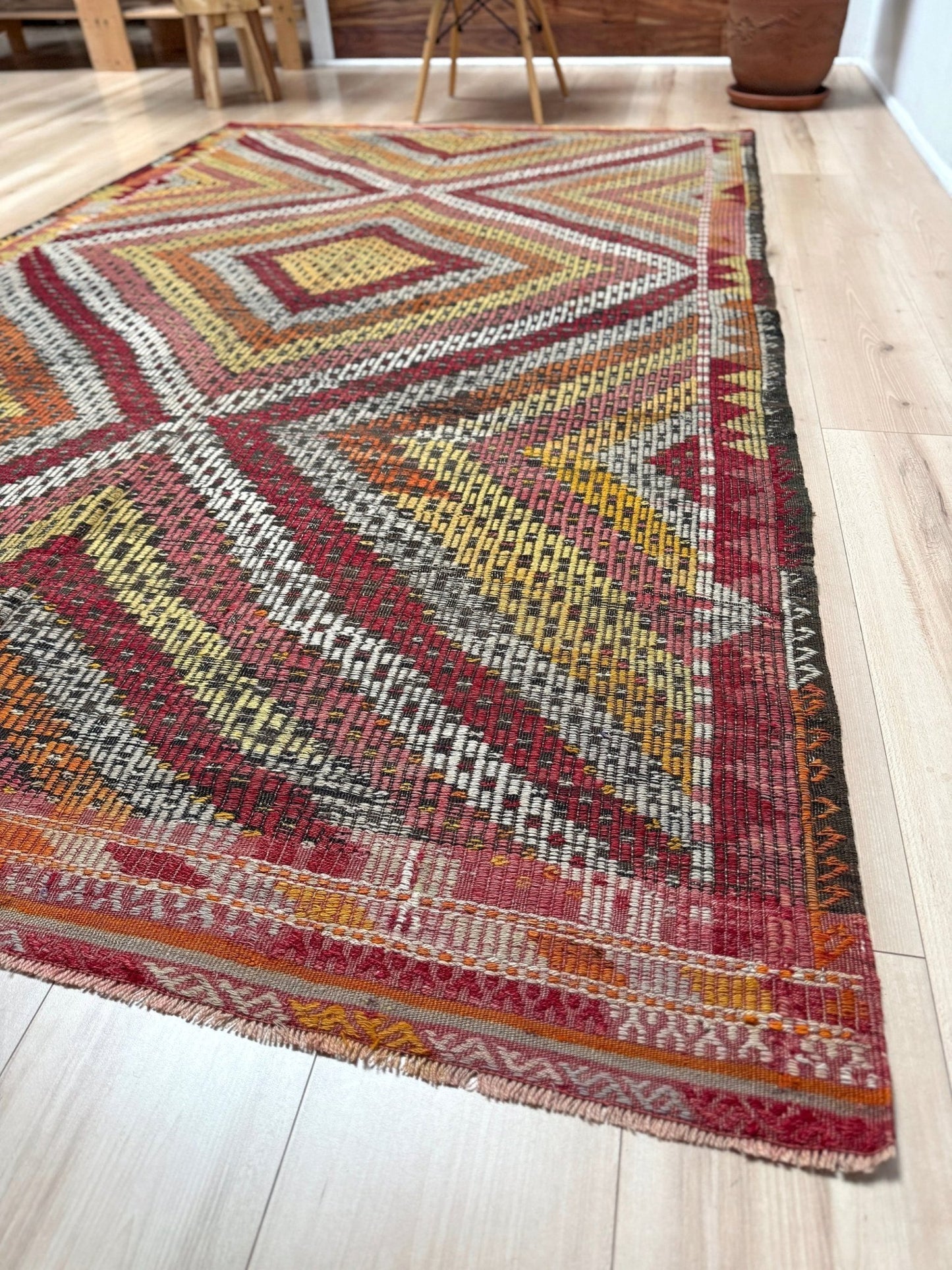 A kilim area rug with a geometric diamond pattern in red, yellow, orange, brown, and white, shown laid out on a light wood floor.  6x9 Vintage Turkish cicim kilim rug with bold diamond pattern, handmade Turkish embroidered wool flatweave from Indigo Rugs Burlingame Bay Area.