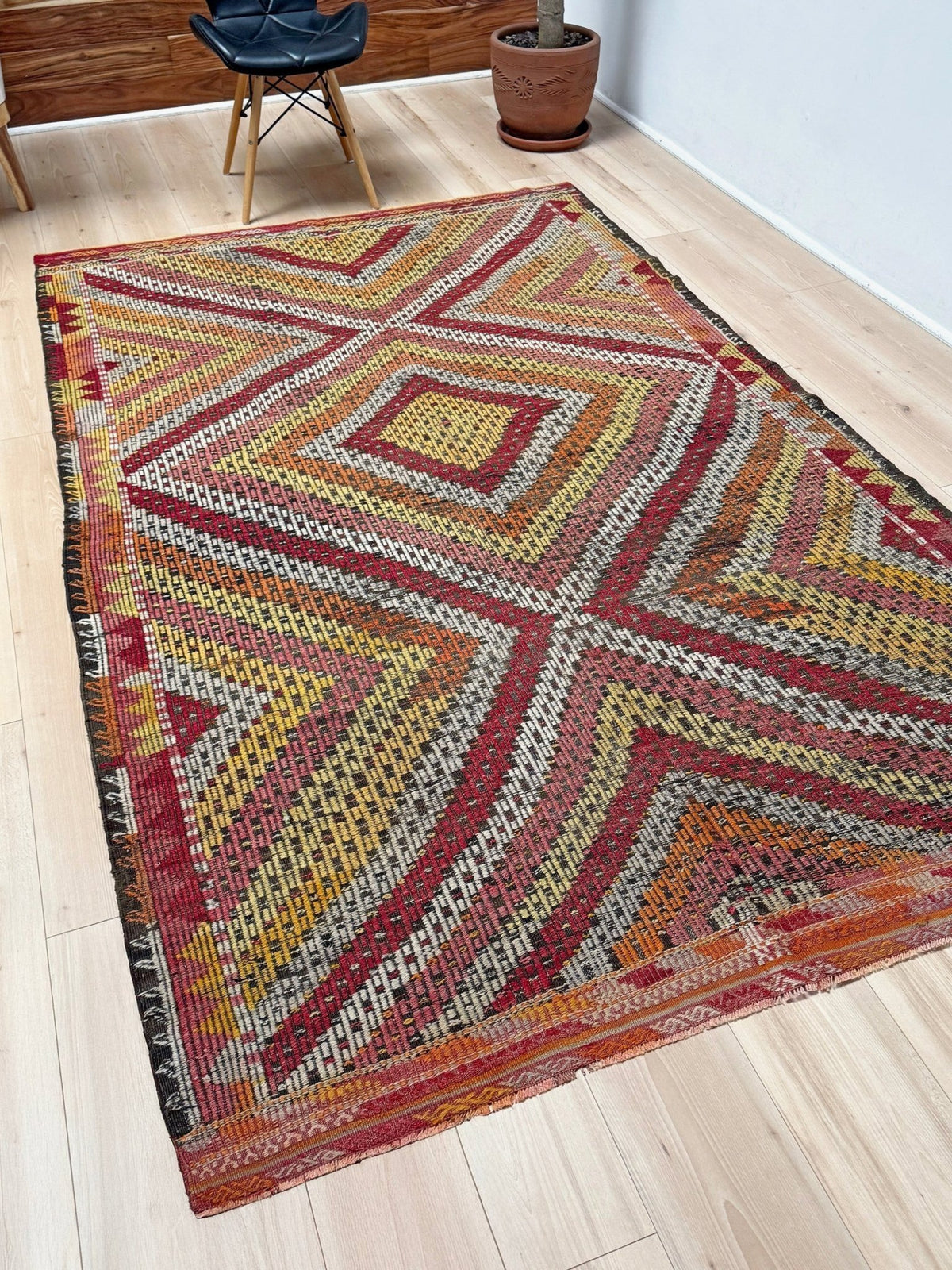 A rectangular woven area rug with a bold geometric diamond and cross pattern in red, orange, yellow, beige, and black, shown on a light wood floor in a room setting.  6x9 Vintage Turkish cicim kilim rug with bold diamond pattern, handmade Turkish embroidered wool flatweave from Indigo Rugs Burlingame Bay Area.