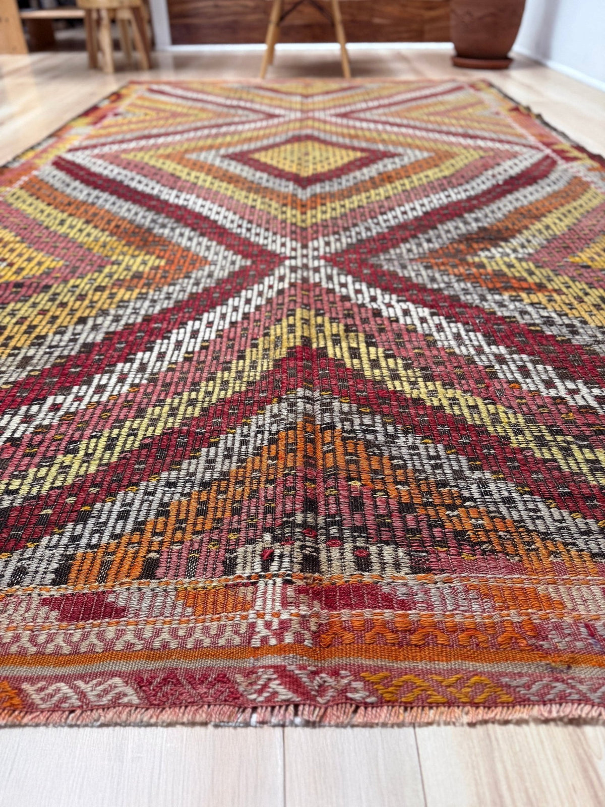 A kilim-style area rug with a geometric diamond and chevron pattern in red, orange, yellow, black, and beige, shown on a light wood floor in a room setting.  6x9 Vintage Turkish cicim kilim rug with bold diamond pattern, handmade Turkish embroidered wool flatweave from Indigo Rugs Burlingame Bay Area.