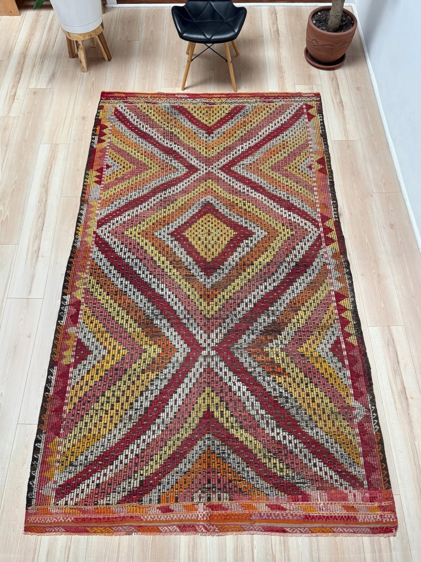 A rectangular vintage-style kilim rug with a geometric diamond and chevron pattern in red, orange, yellow, and beige, laid out on a light wood floor in a modern room.  6x9 Vintage Turkish cicim kilim rug with bold diamond pattern, handmade Turkish embroidered wool flatweave from Indigo Rugs Burlingame Bay Area.