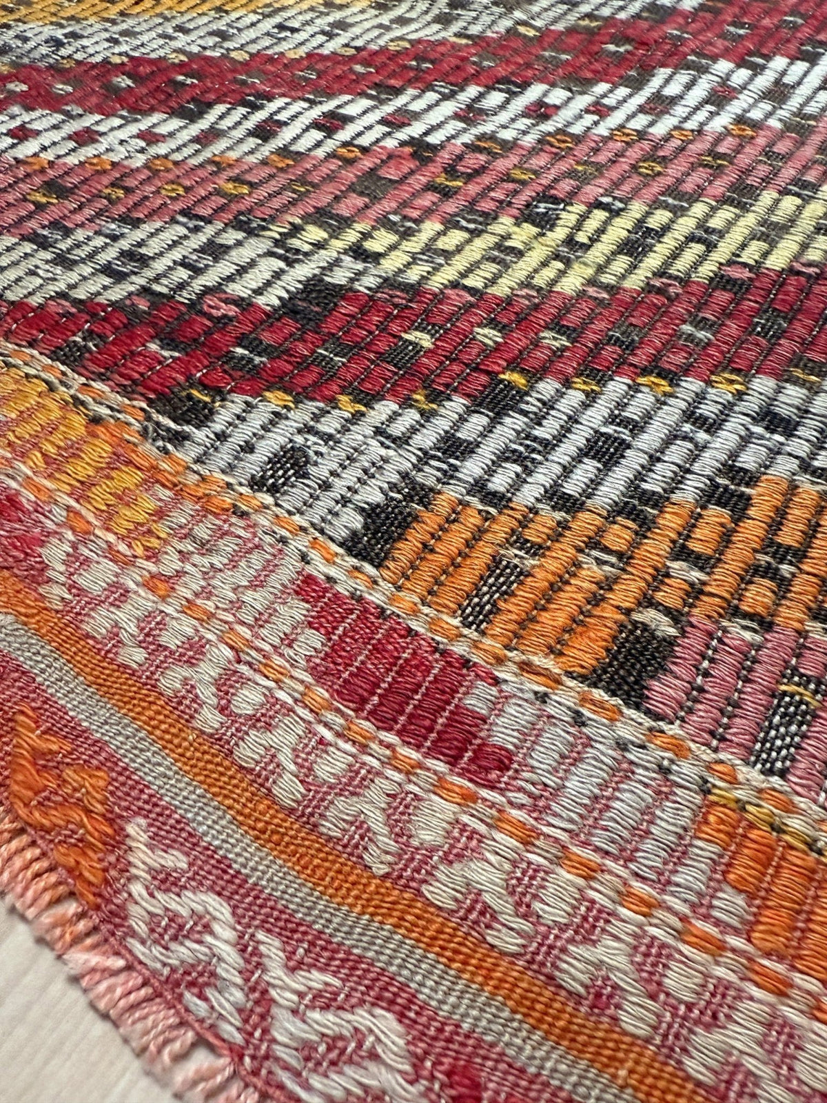 Close-up of a woven textile with a multicolor geometric pattern in red, orange, pink, black, white, and yellow, showing detailed thread texture and fringe along one edge.  6x9 Vintage Turkish cicim kilim rug with bold diamond pattern, handmade Turkish embroidered wool flatweave from Indigo Rugs Burlingame Bay Area.