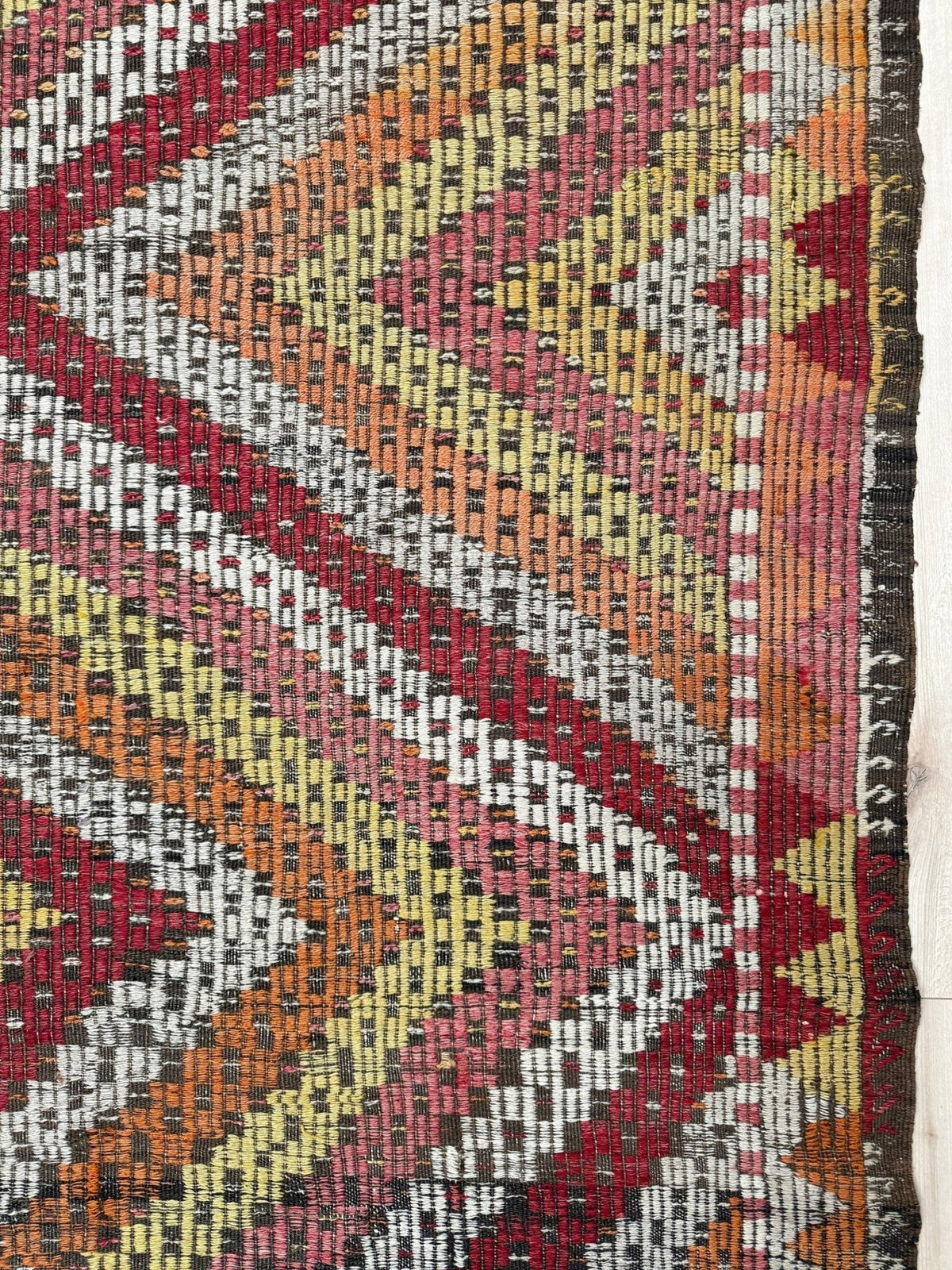 A close-up of a woven kilim rug showing a geometric chevron pattern in red, orange, yellow, pink, and gray with a dark fringe on the right side.  6x9 Vintage Turkish cicim kilim rug with bold diamond pattern, handmade Turkish embroidered wool flatweave from Indigo Rugs Burlingame Bay Area.