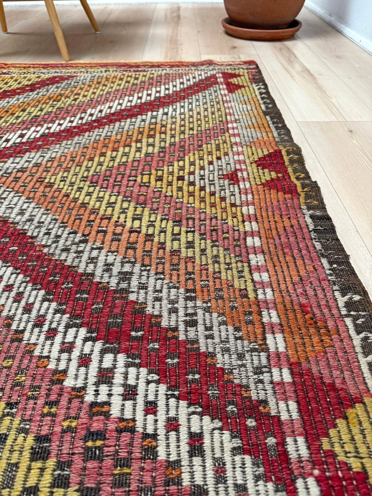 A close-up of a woven area rug with a geometric tribal pattern in red, orange, yellow, cream, and dark brown, laid out on a light wood floor.  6x9 Vintage Turkish cicim kilim rug with bold diamond pattern, handmade Turkish embroidered wool flatweave from Indigo Rugs Burlingame Bay Area.
