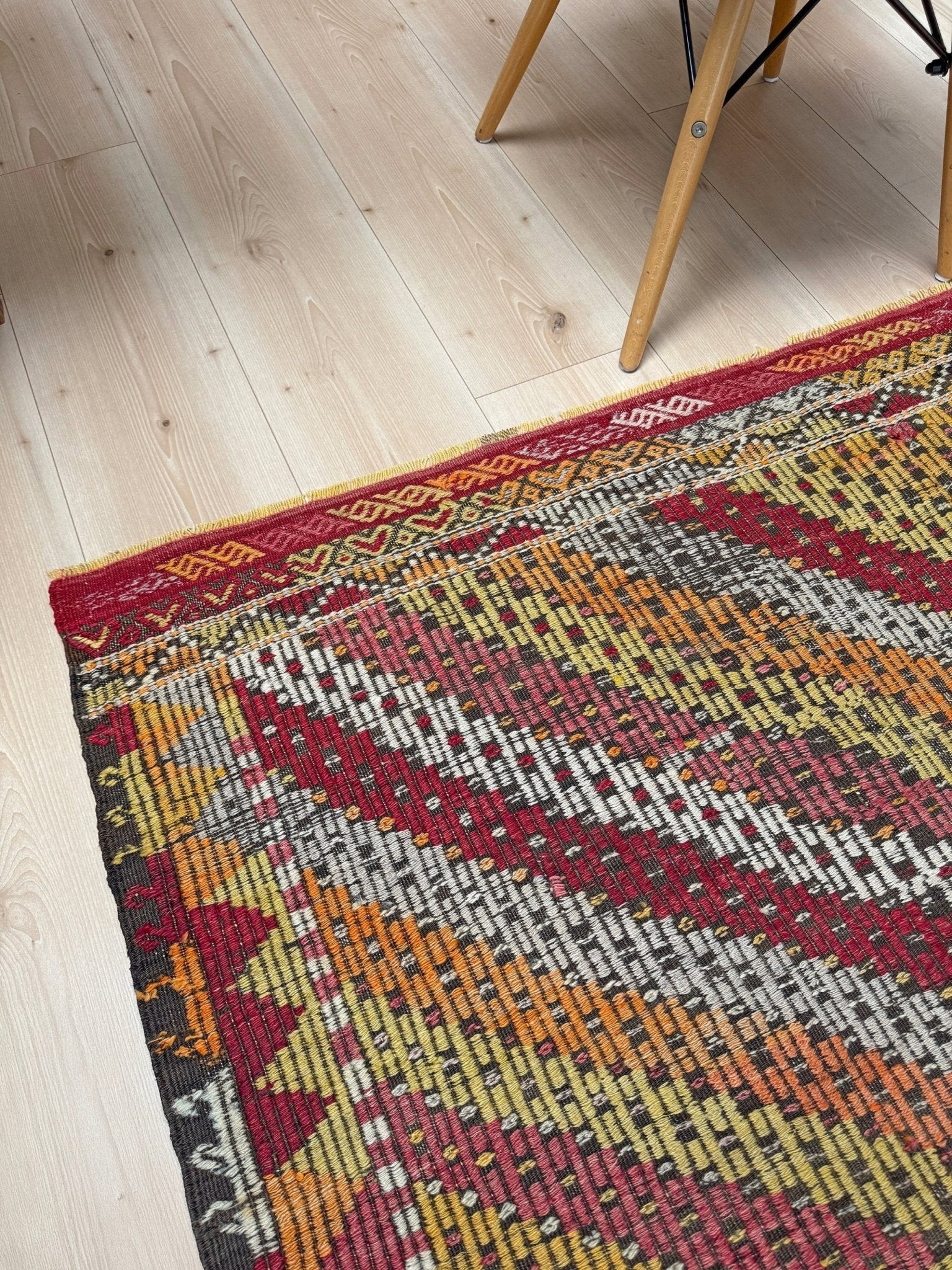 A close-up of a colorful woven area rug with geometric patterns, shown on a light wood floor next to the legs of a wooden chair.  6x9 Vintage Turkish cicim kilim rug with bold diamond pattern, handmade Turkish embroidered wool flatweave from Indigo Rugs Burlingame Bay Area.