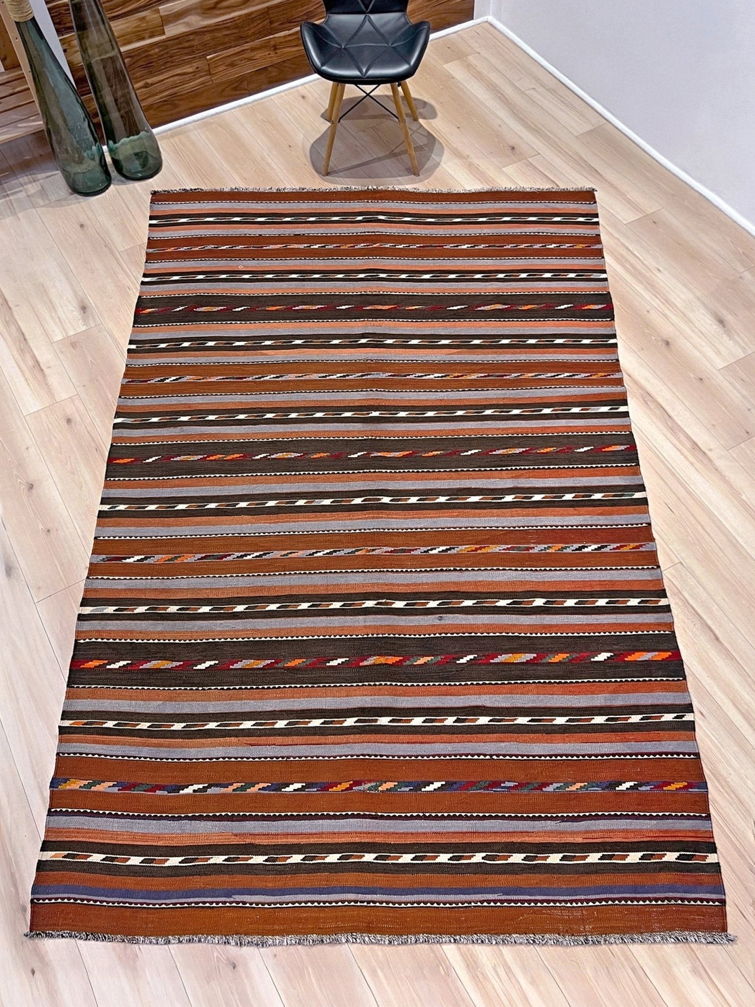 Striped rug on a wooden floor with a chair in the background. Vintage Caucasian kilim rug with horizontal tribal stripes in rust, brown, gray, and ivory tones, handwoven wool flatweave rug 6x9